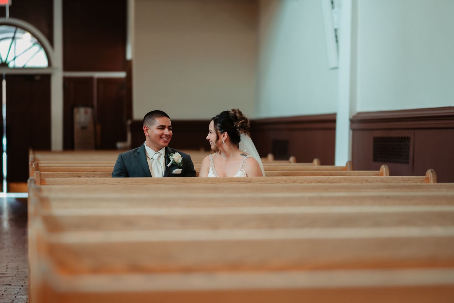 A couple kisses in a church pew during their wedding ceremony while family members watch from nearby pews.