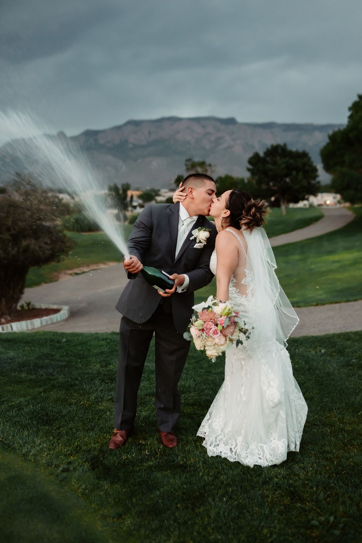Playful newlyweds spraying champagne on grassy hill with mountains in background.