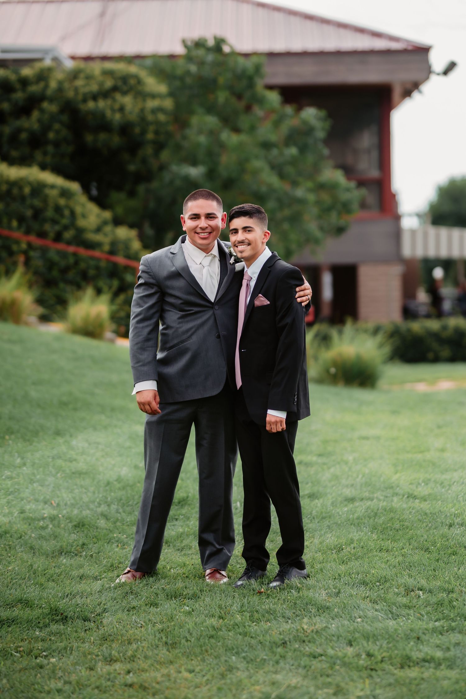 Two young attendants in formal suits smile and embrace each other on a grassy hill during a wedding.