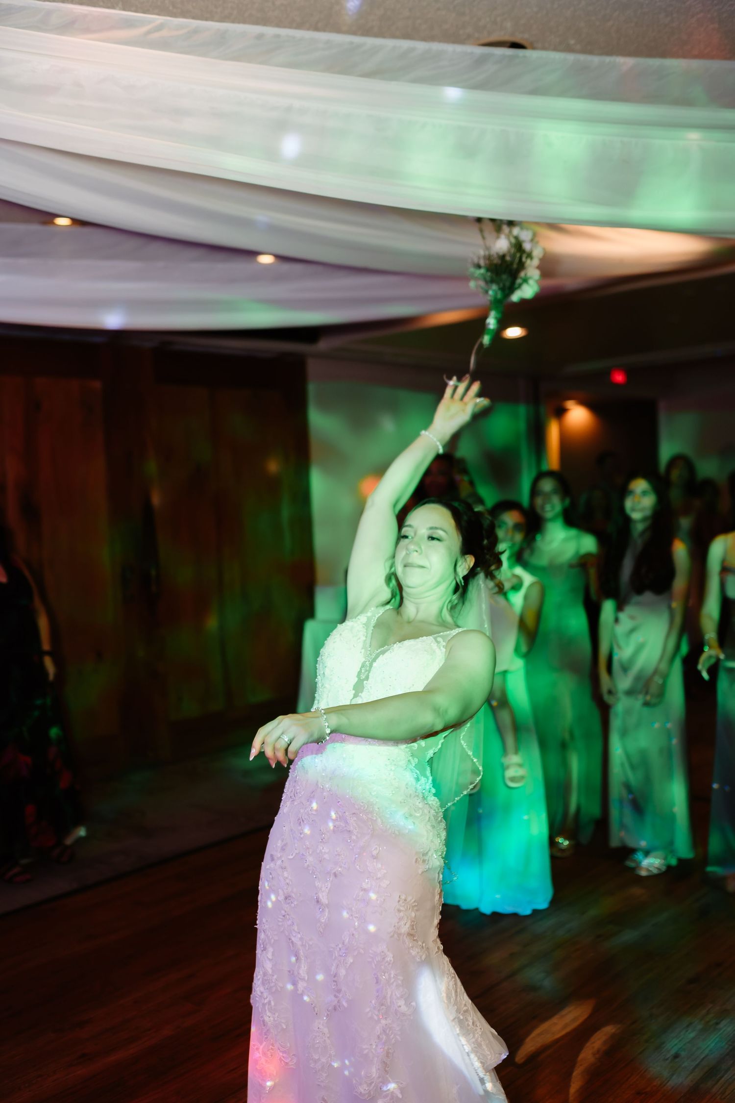 A bride dances joyfully under green lighting and white ceiling draping at an indoor wedding reception.