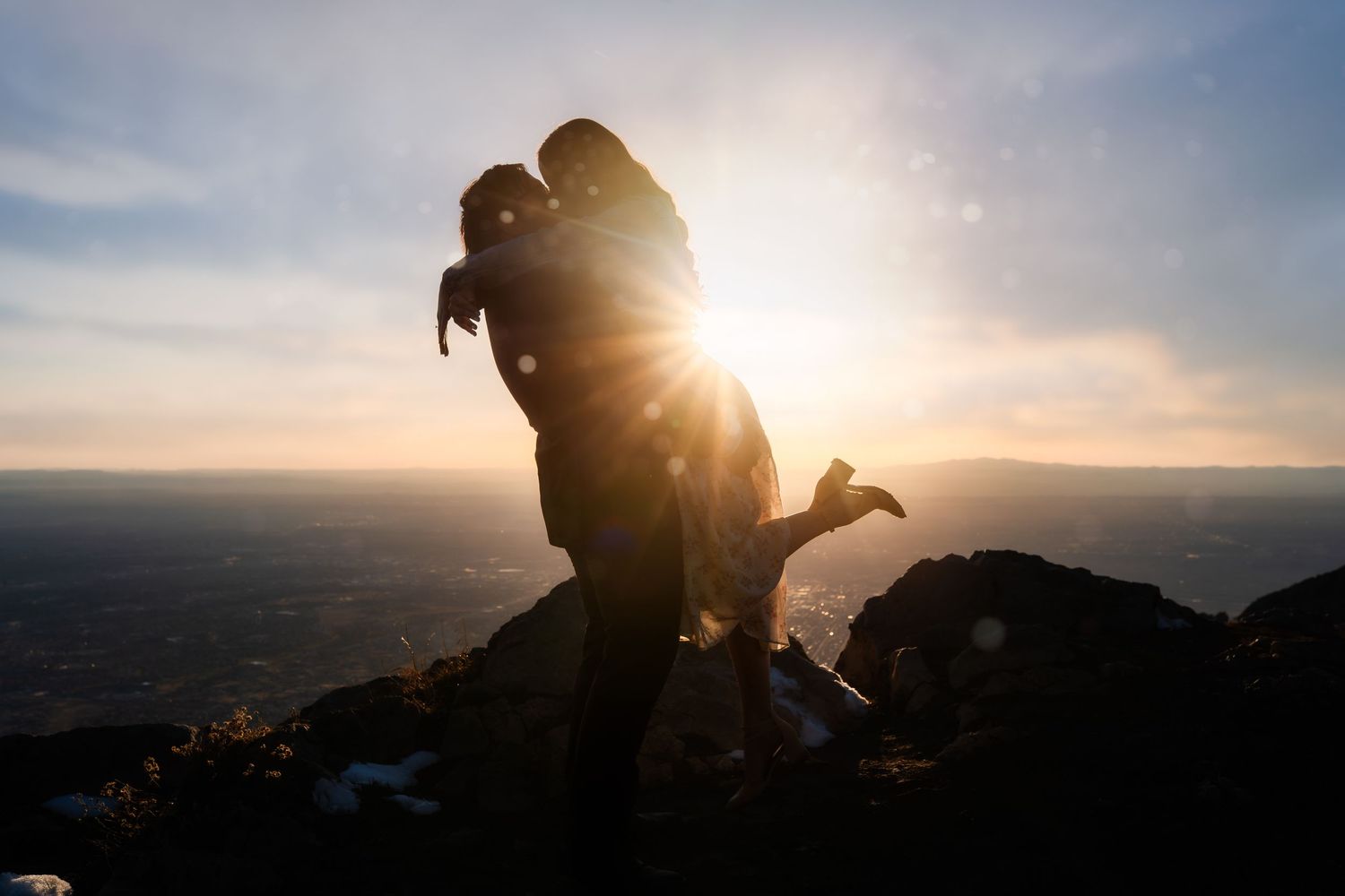 Silhouetted couple sharing romantic kiss at sunset on mountain overlook with sun flare behind them.