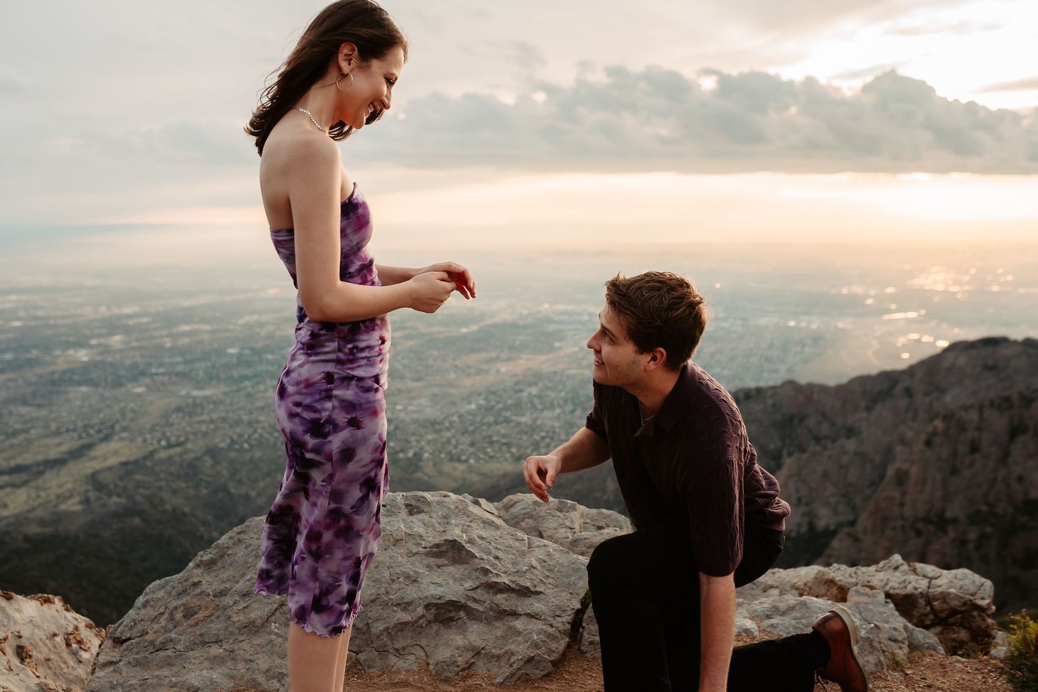 A couple in casual attire stands on top of a mountain peak at sunset during what appears to be a proposal moment.