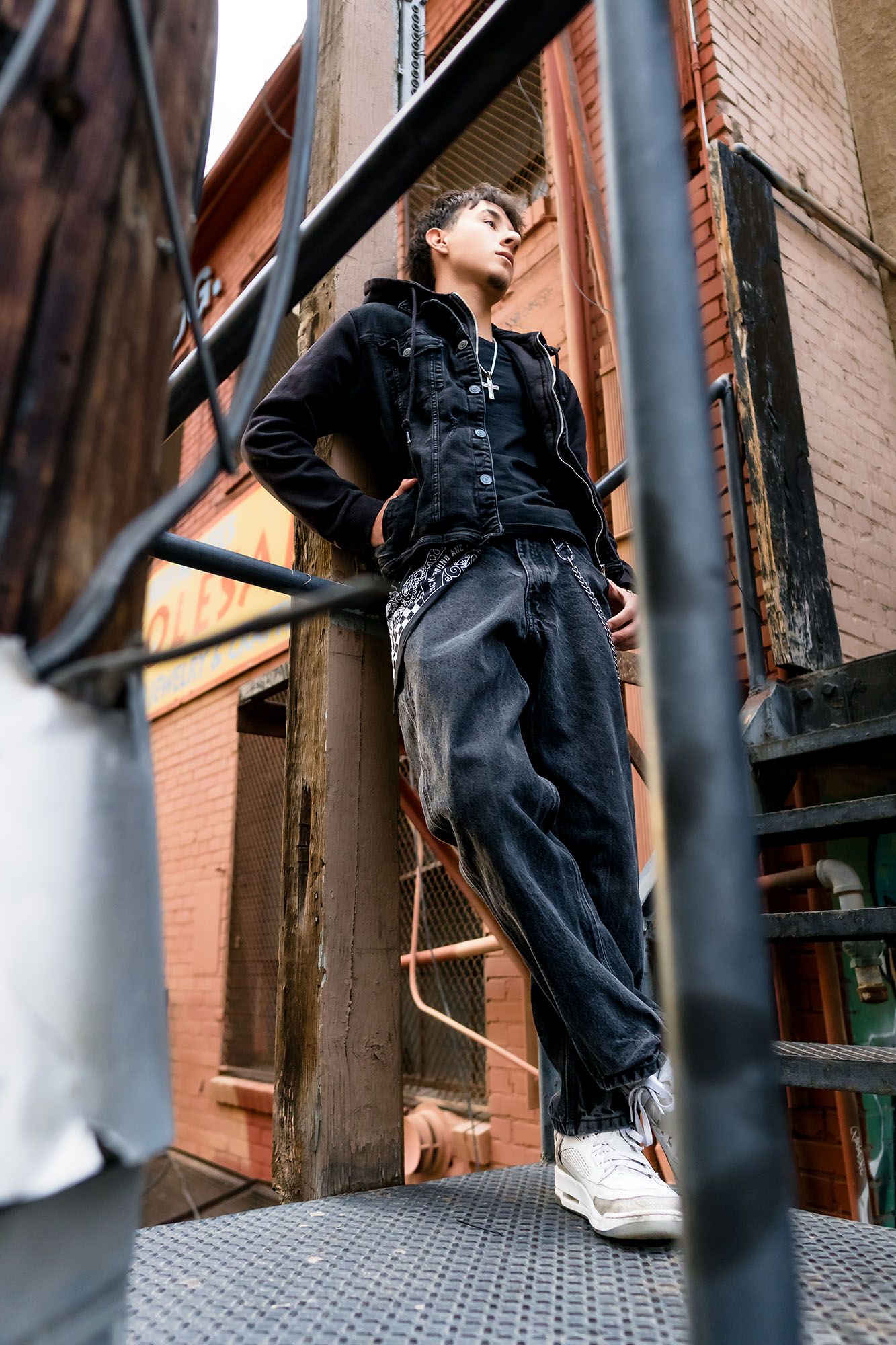 Young person in black denim jacket and dark jeans poses on urban fire escape stairway.