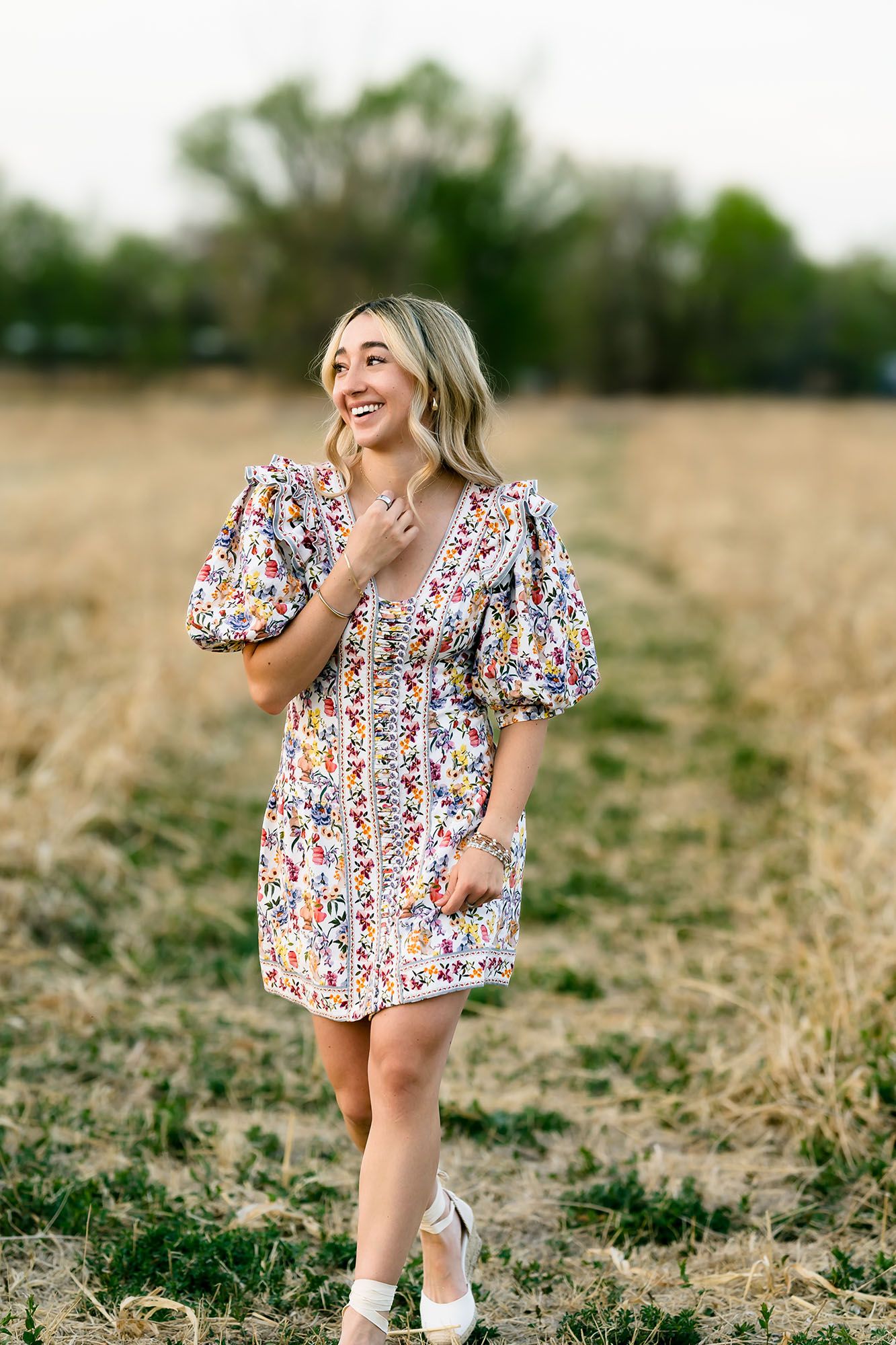 Smiling young woman in floral dress walking through golden field during outdoor photo session.