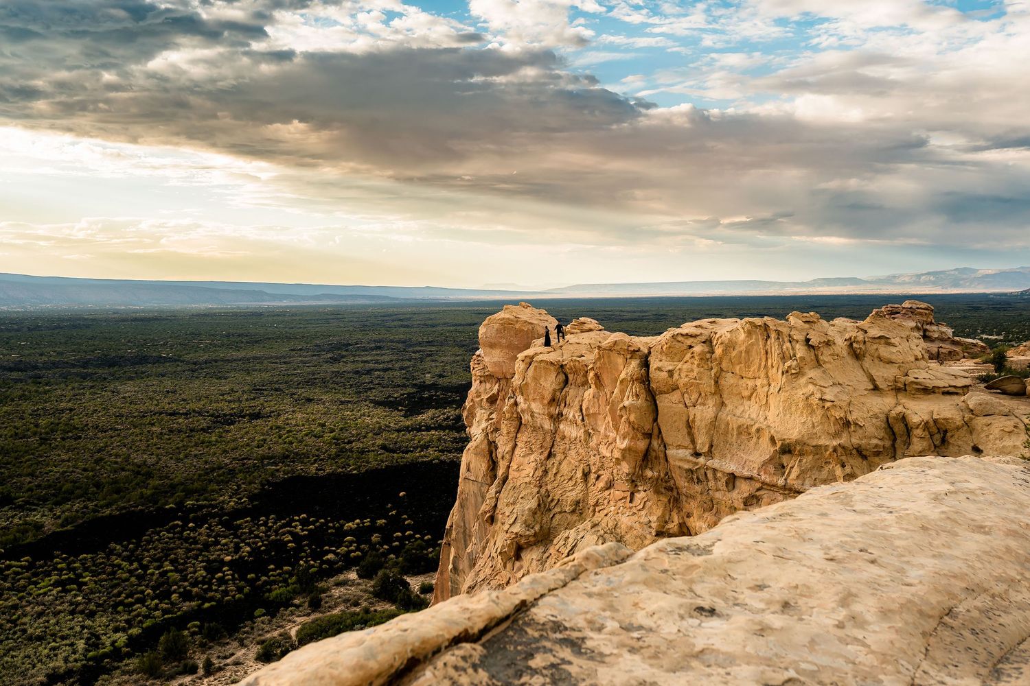 Sweeping aerial view of a vast desert landscape with dramatic sandstone cliffs under a cloudy sunset sky.