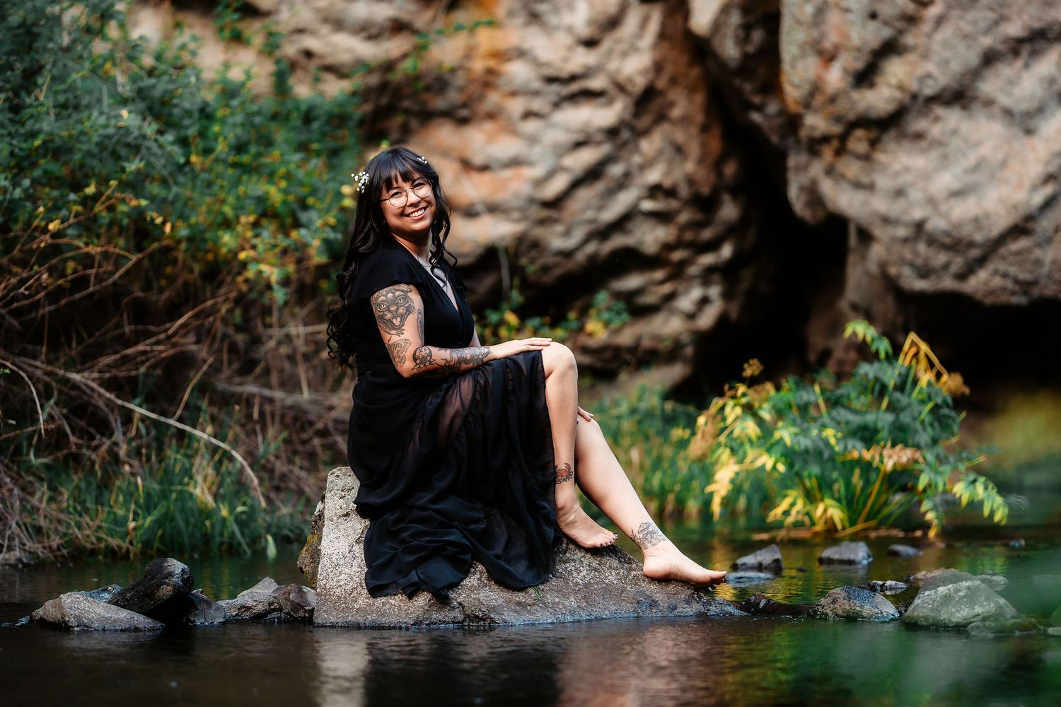 A tattooed woman in a black dress sits barefoot on rocks by a serene creek in nature.