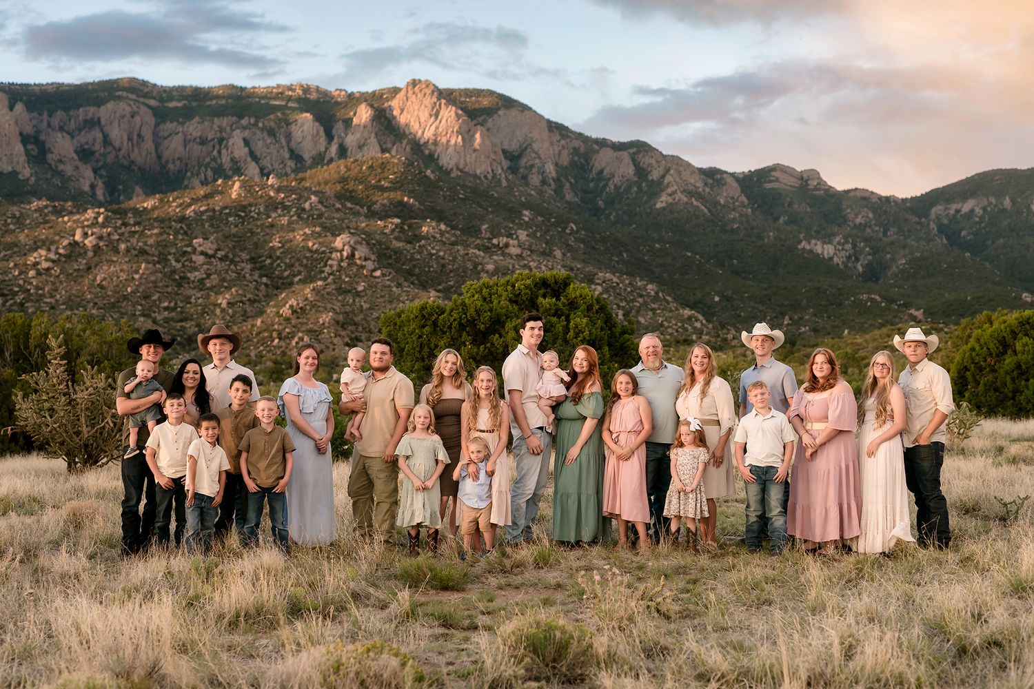 Large family portrait outdoors with mountain backdrop at golden hour, adults and children in coordinated earth-tone outfits.