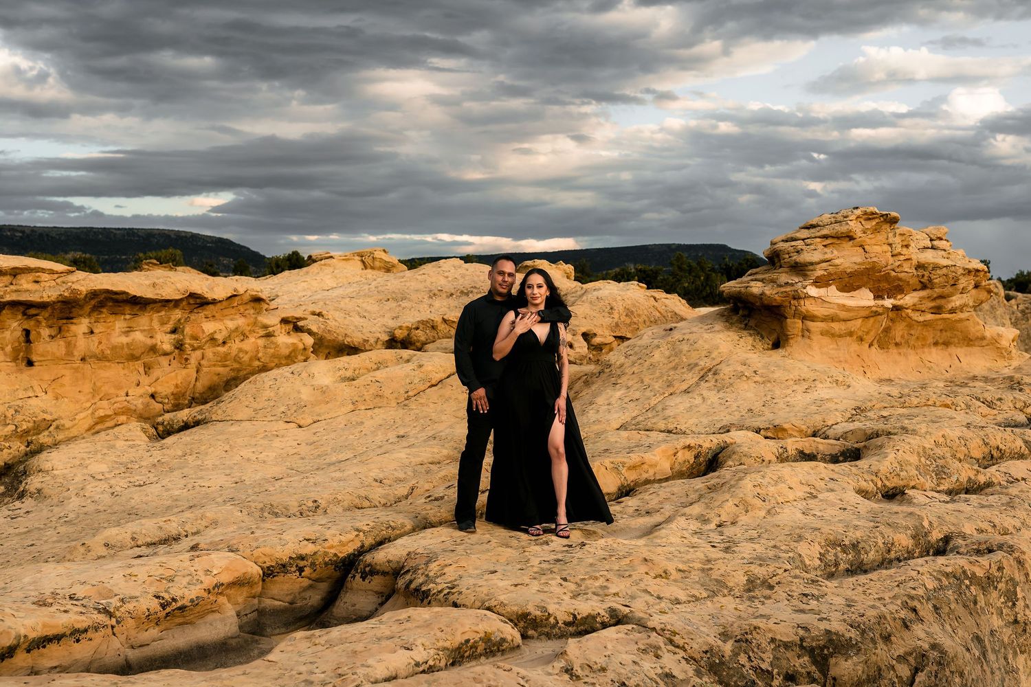Elegant couple in black formal wear standing on golden sandstone rocks with moody storm clouds above.