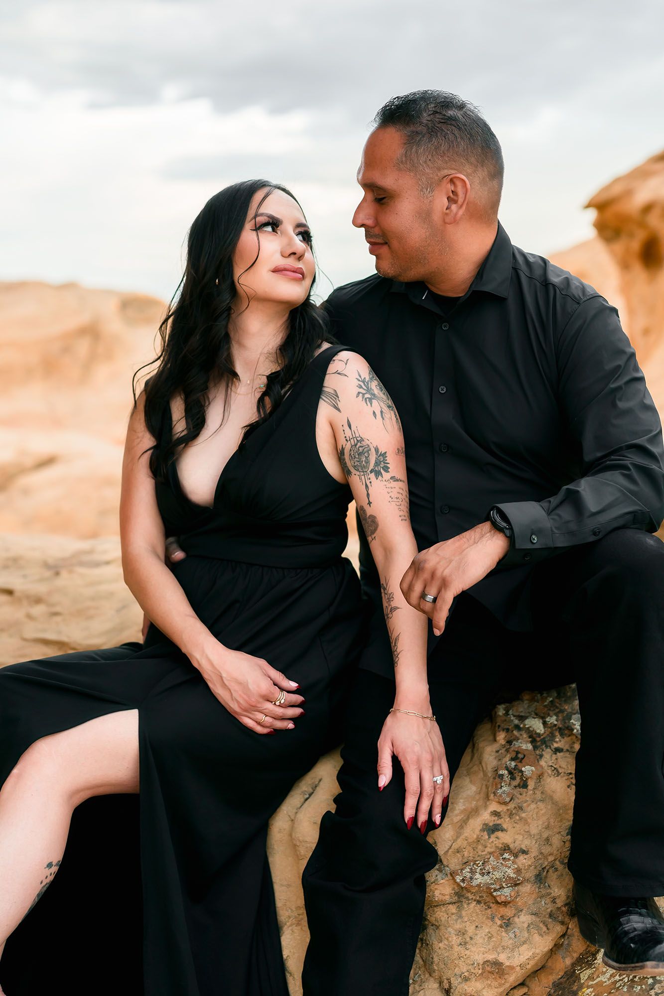 Couple in matching black outfits sharing a romantic moment seated on rocks in a desert landscape.