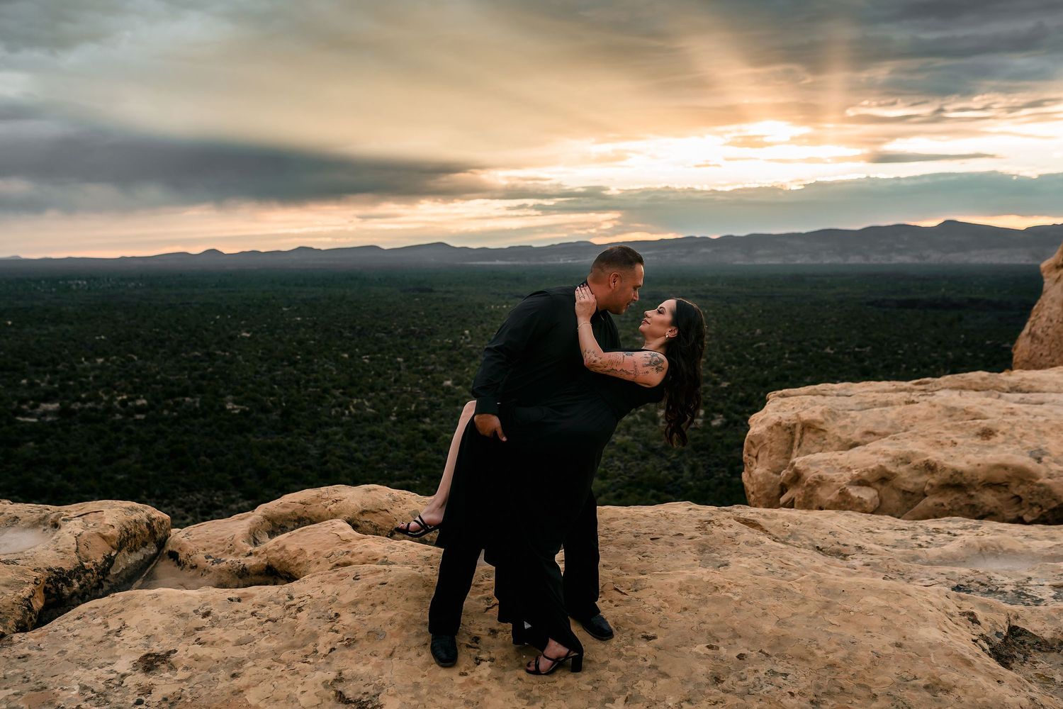 Couple in black sharing a romantic dip kiss on rocky cliff with stunning sunset panorama.