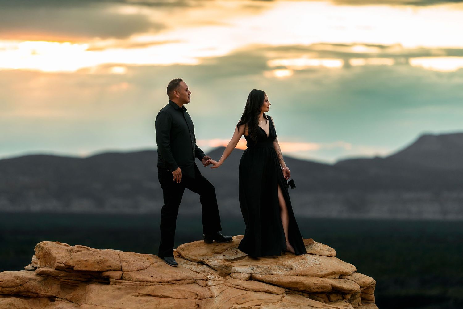 Couple in black holding hands on rocky cliff edge with golden sunset clouds and mountain landscape behind.