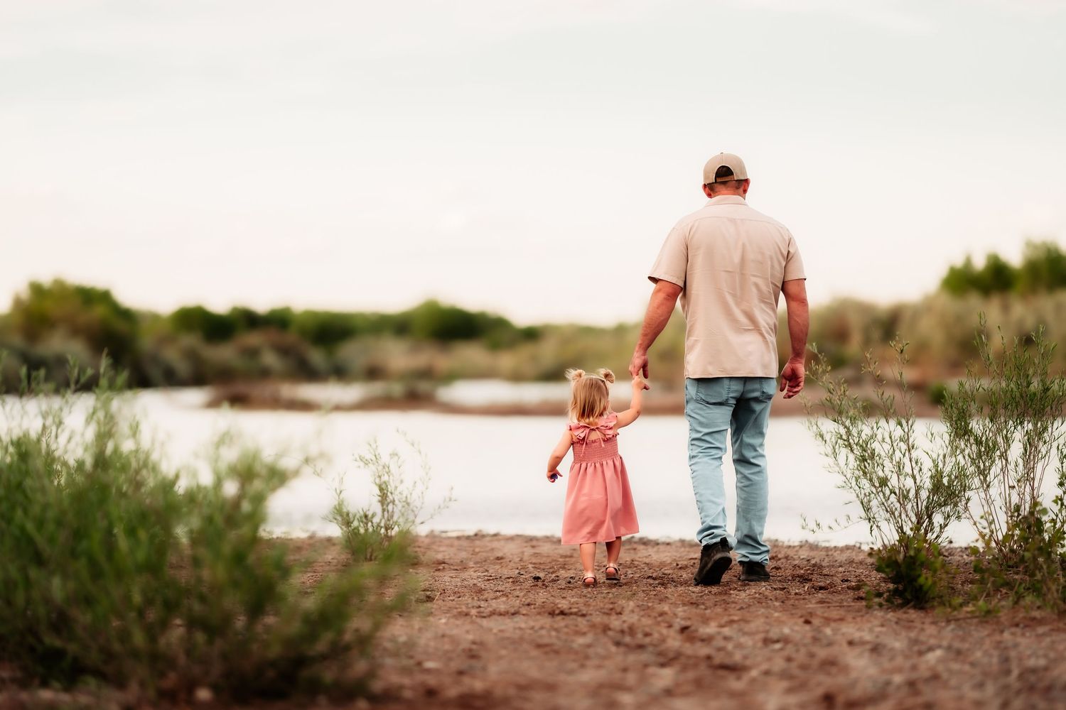 A person in jeans and their small companion in a pink dress walk hand in hand along a riverbank path at sunset.