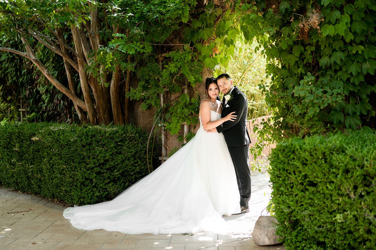 Bride and groom embrace in a lush garden setting, bride wearing a flowing white gown with long train.
