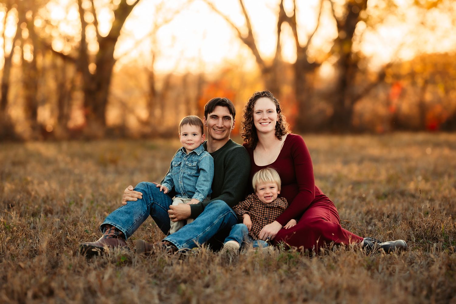 Family of four sitting in autumn field at golden hour, parents with two young boys smiling outdoors.
