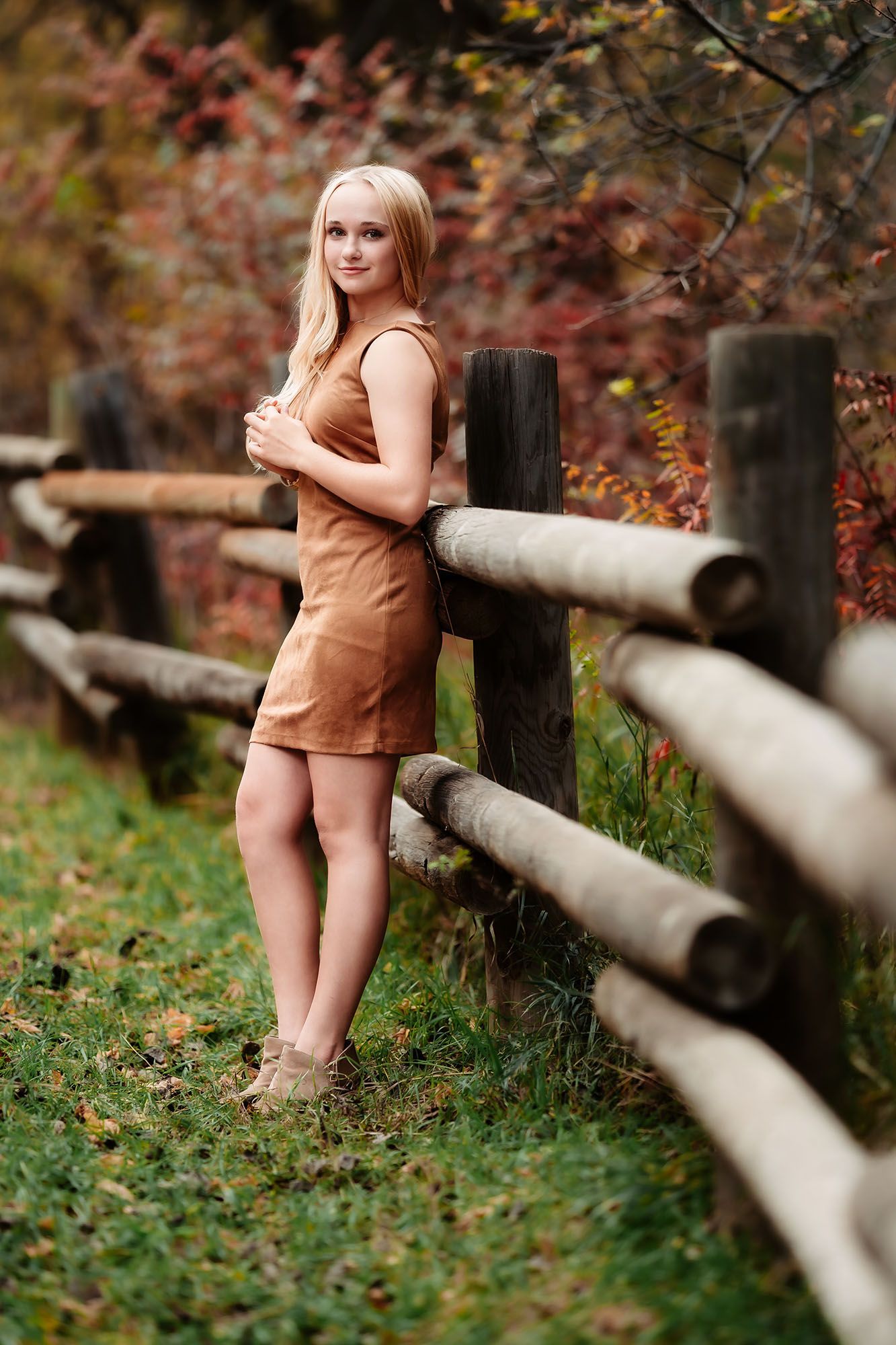 Blonde woman in brown mini dress leaning against rustic wooden fence in autumn forest setting.