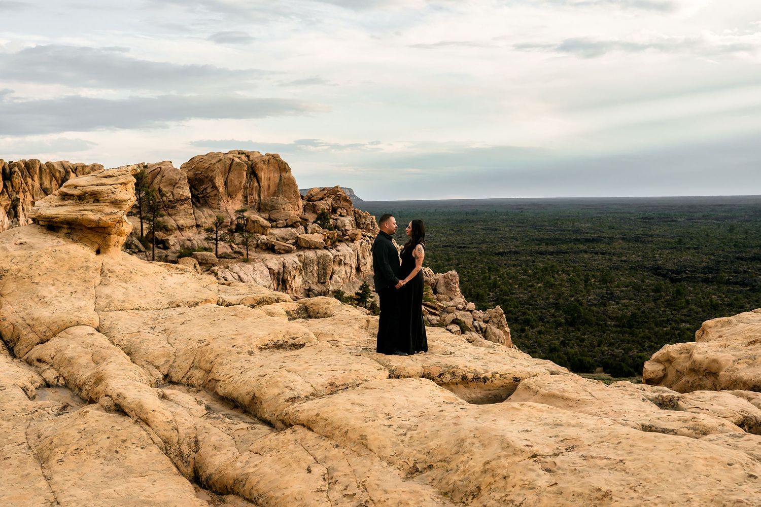 Couple in black attire standing on rocky desert cliff overlooking vast green landscape under cloudy sky.