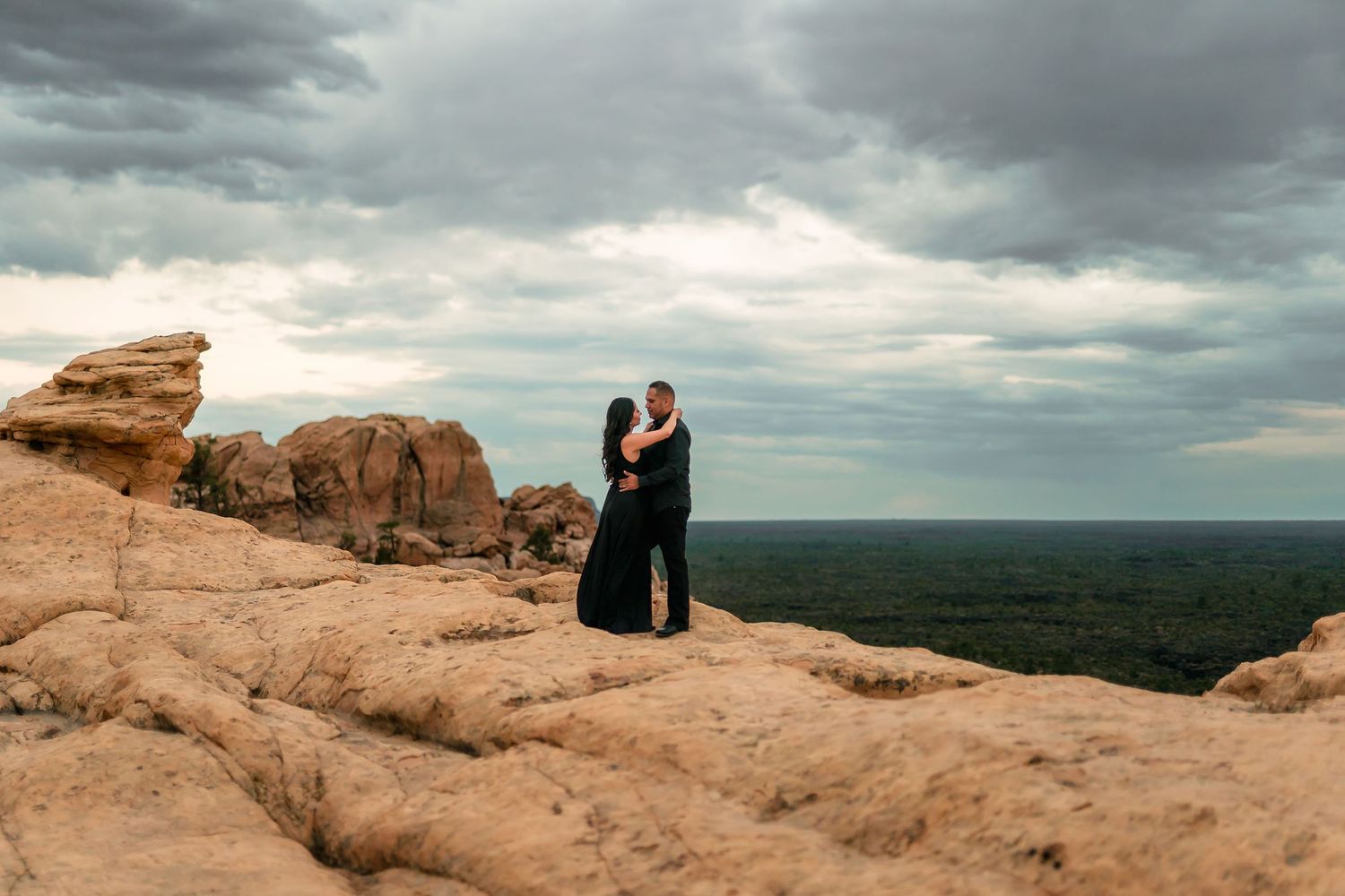 Couple embracing on red rock formation under dramatic stormy sky with vast landscape in background.