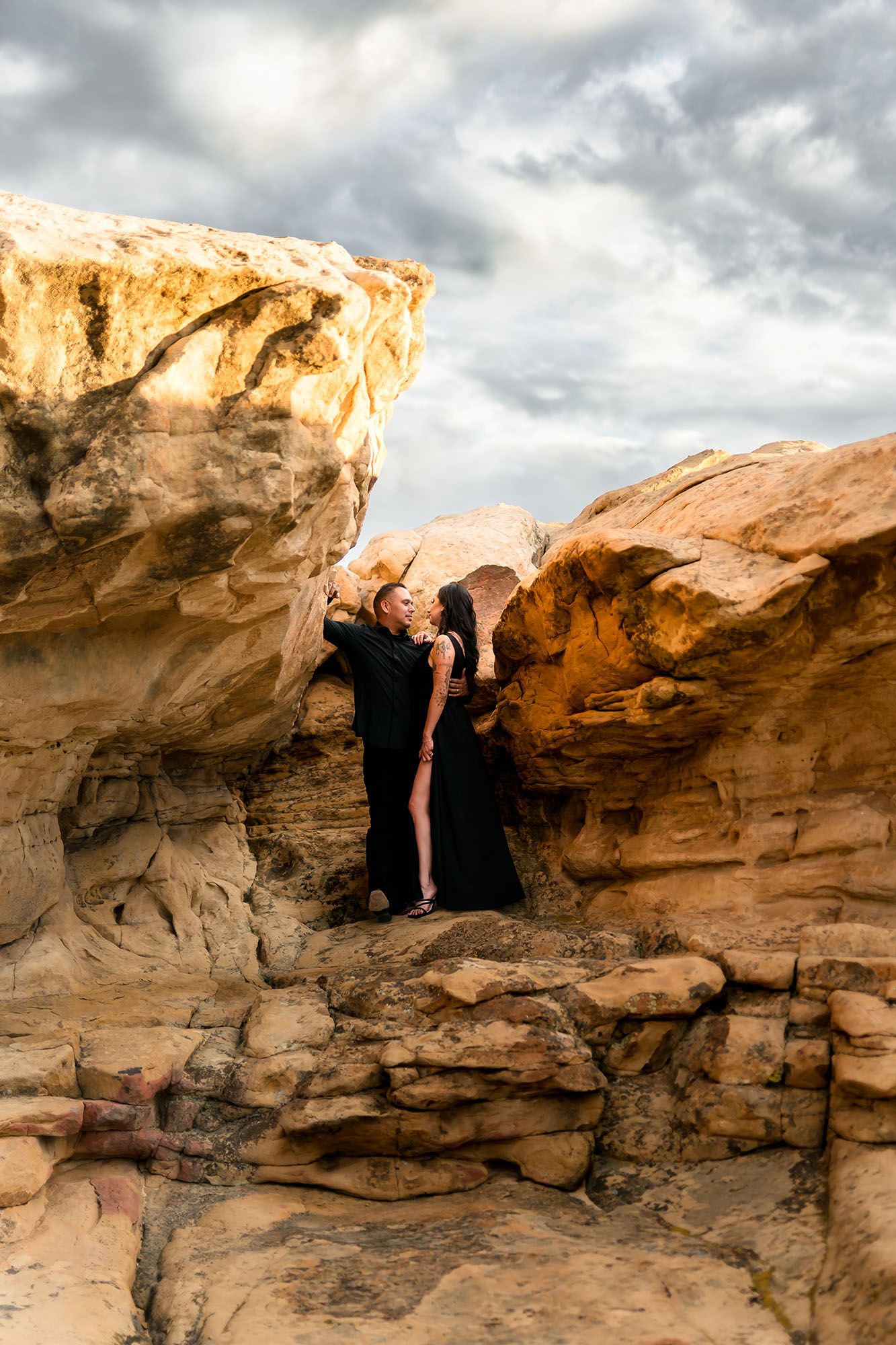 Couple in elegant black attire embracing between dramatic sandstone rock formations under cloudy sky.