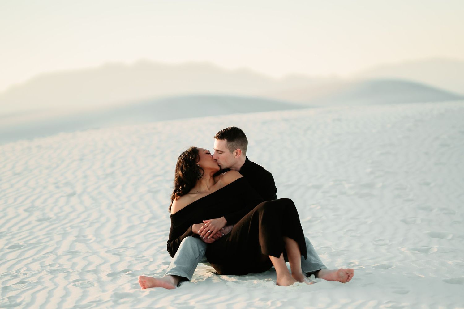 A romantic couple sits intimately together on pristine white sand dunes at dusk.