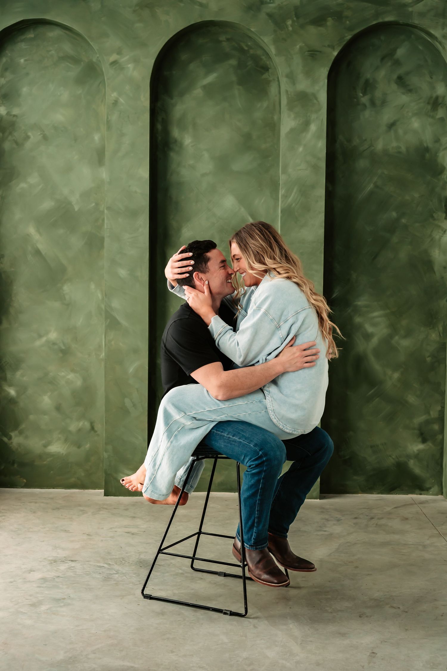 A romantic couple embraces while sitting on a bar stool against a green textured wall with arched alcoves.