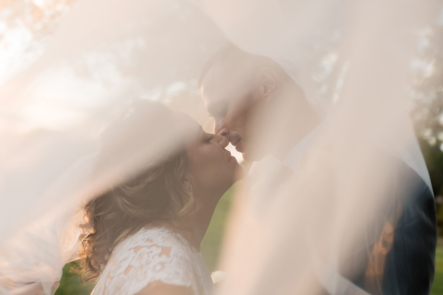 A bride and groom share a romantic kiss beneath a flowing veil at golden hour during their wedding day.