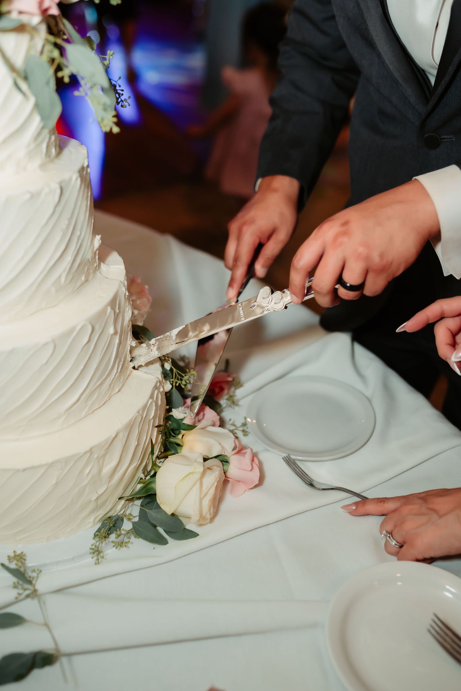 Close-up shot of newlyweds cutting their white tiered wedding cake adorned with roses and greenery.