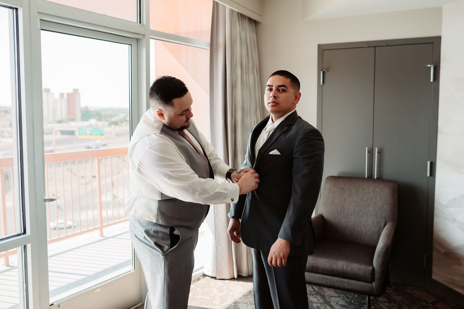 Groomsmen in suits getting ready in bright hotel room with city views.