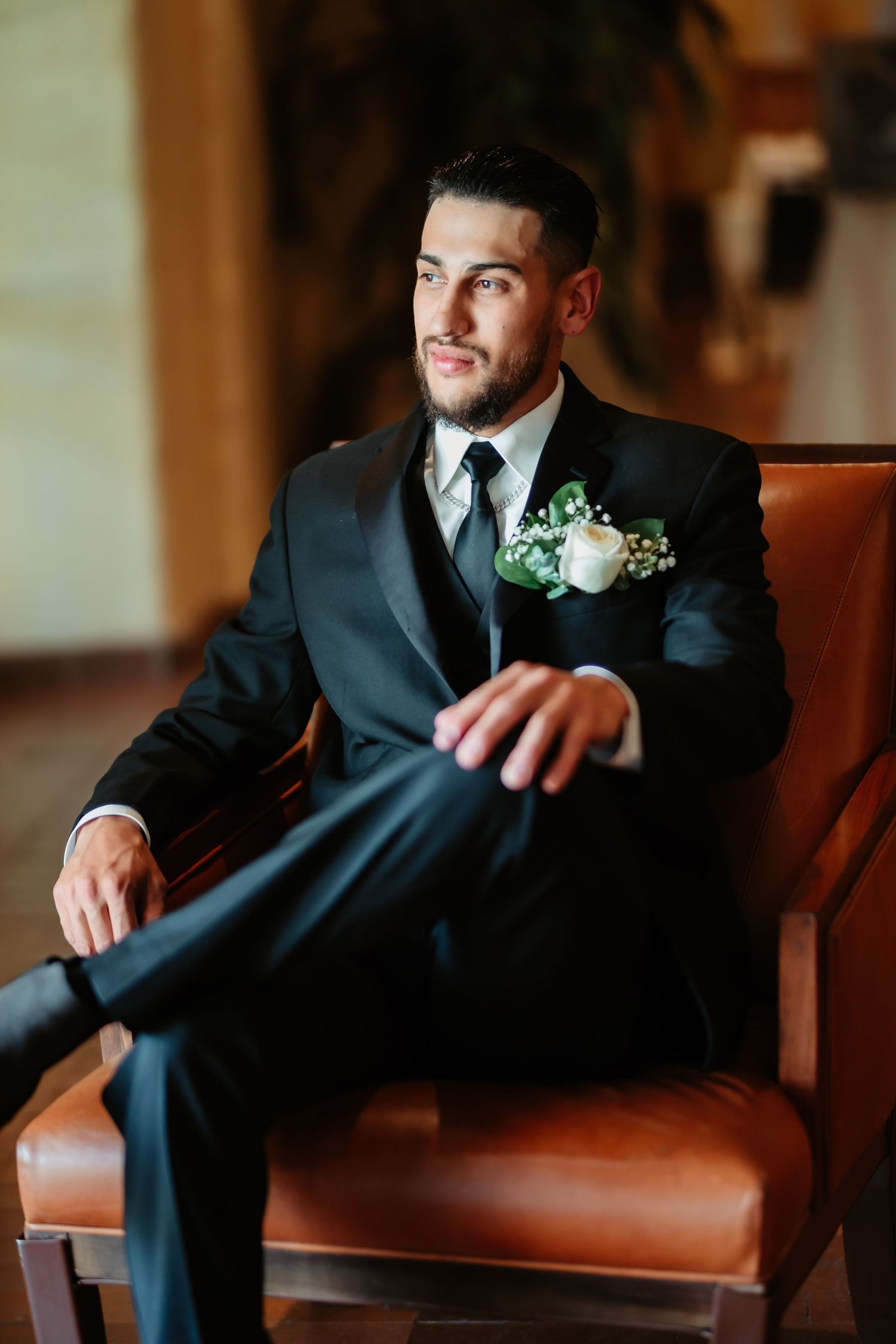 A groom in a black suit sits on a leather bench in various poses during a wedding photoshoot.