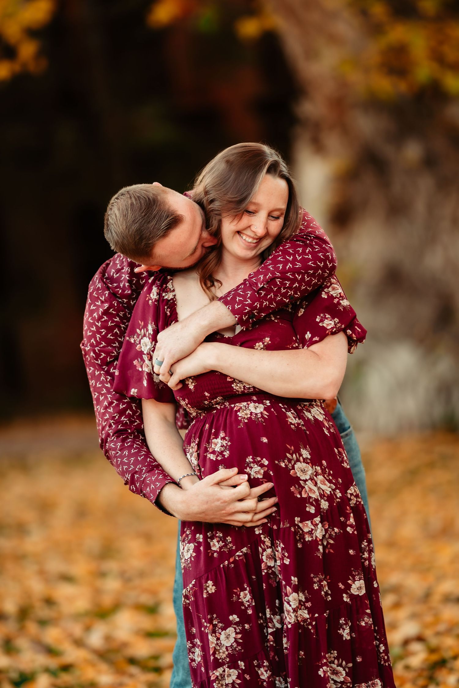 A couple embraces in a romantic autumn setting with fallen leaves and burgundy floral dress.