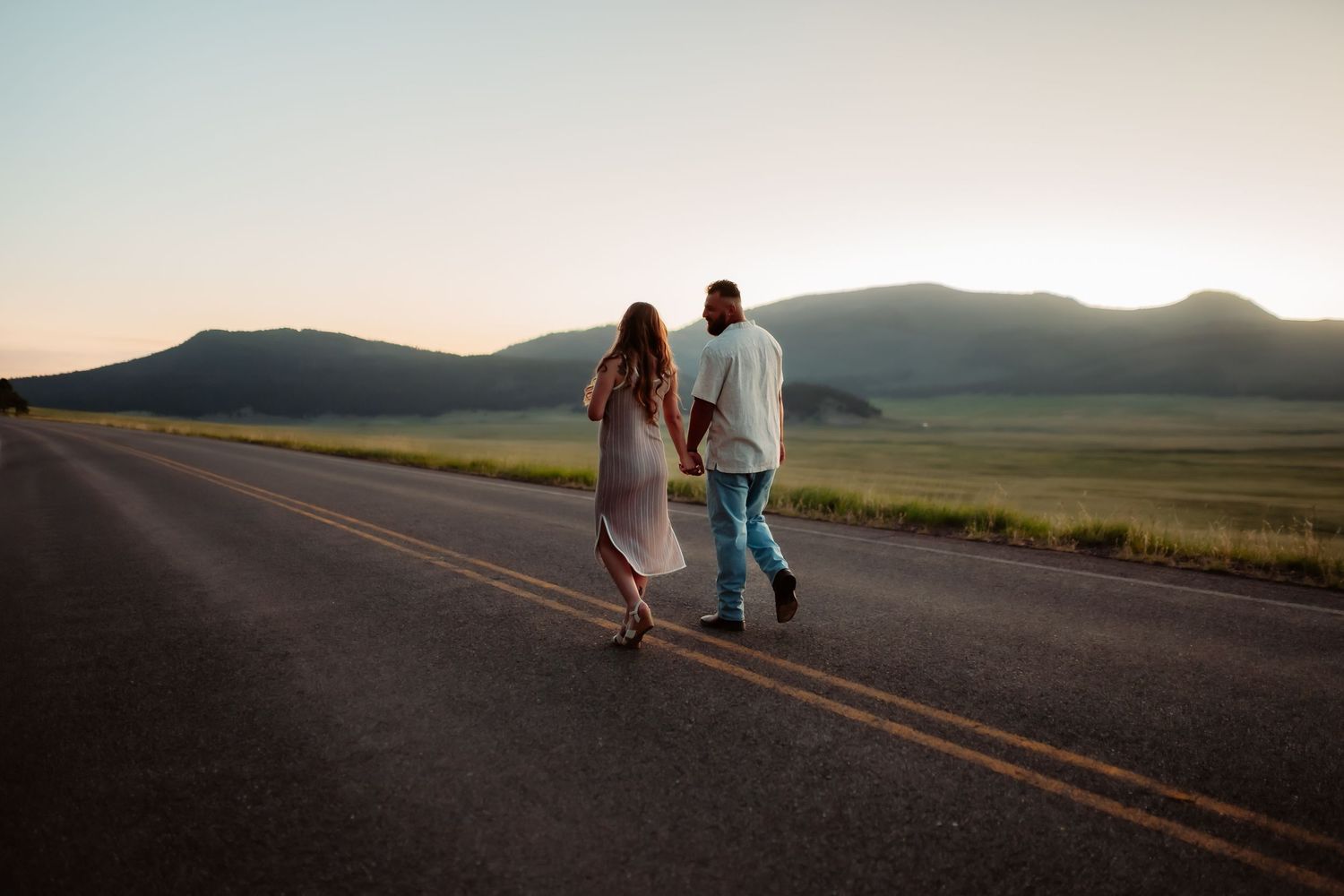 Two people walk hand in hand down an empty rural road at sunset casting long shadows across the pavement.