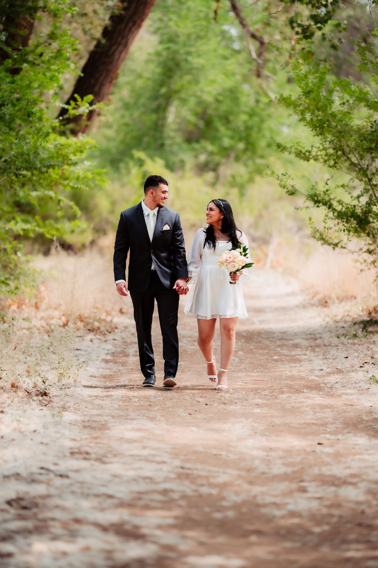 A couple in formal attire walking together on a scenic dirt path surrounded by lush green trees and foliage.