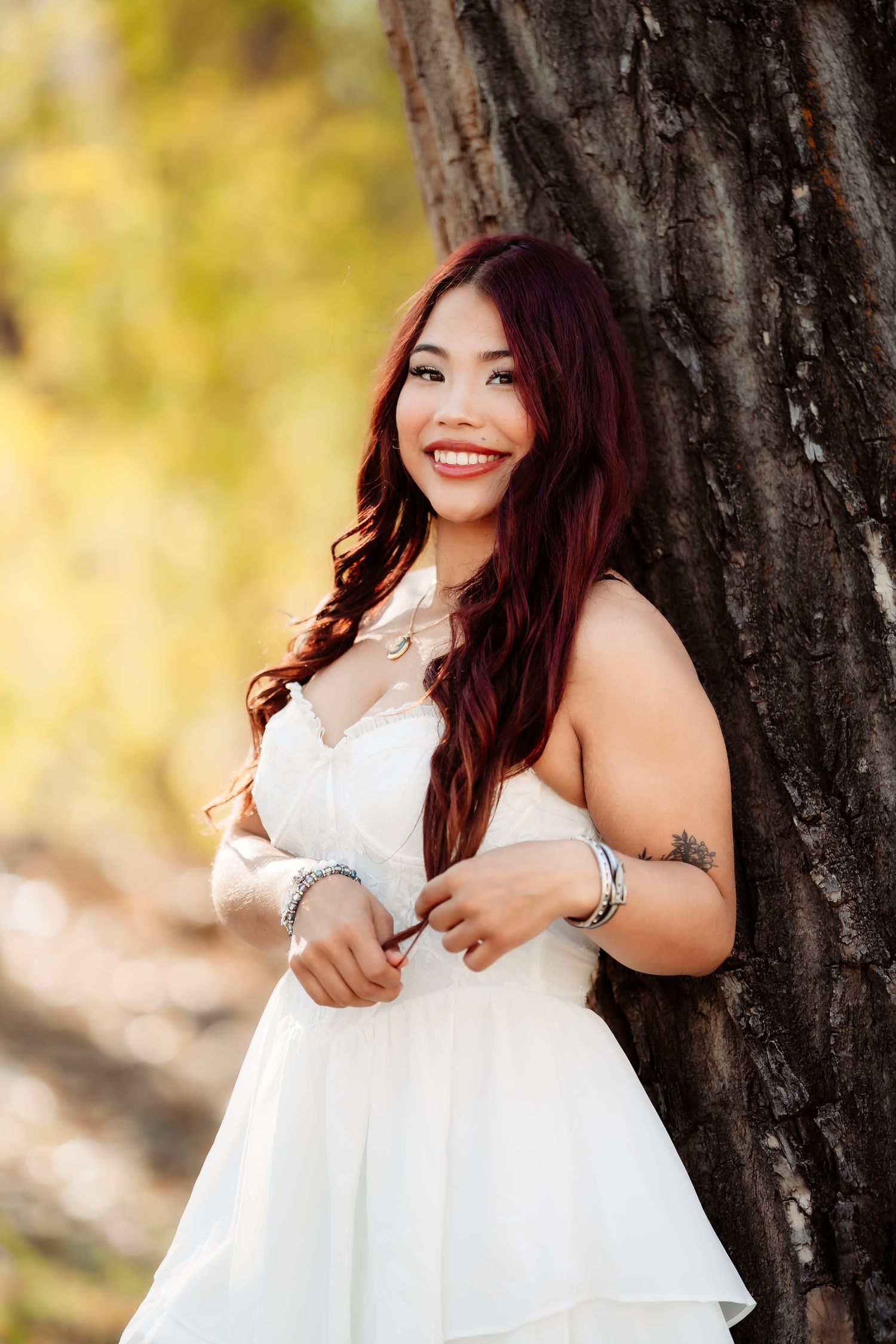Woman in flowing white dress poses against tree trunk in natural outdoor setting.