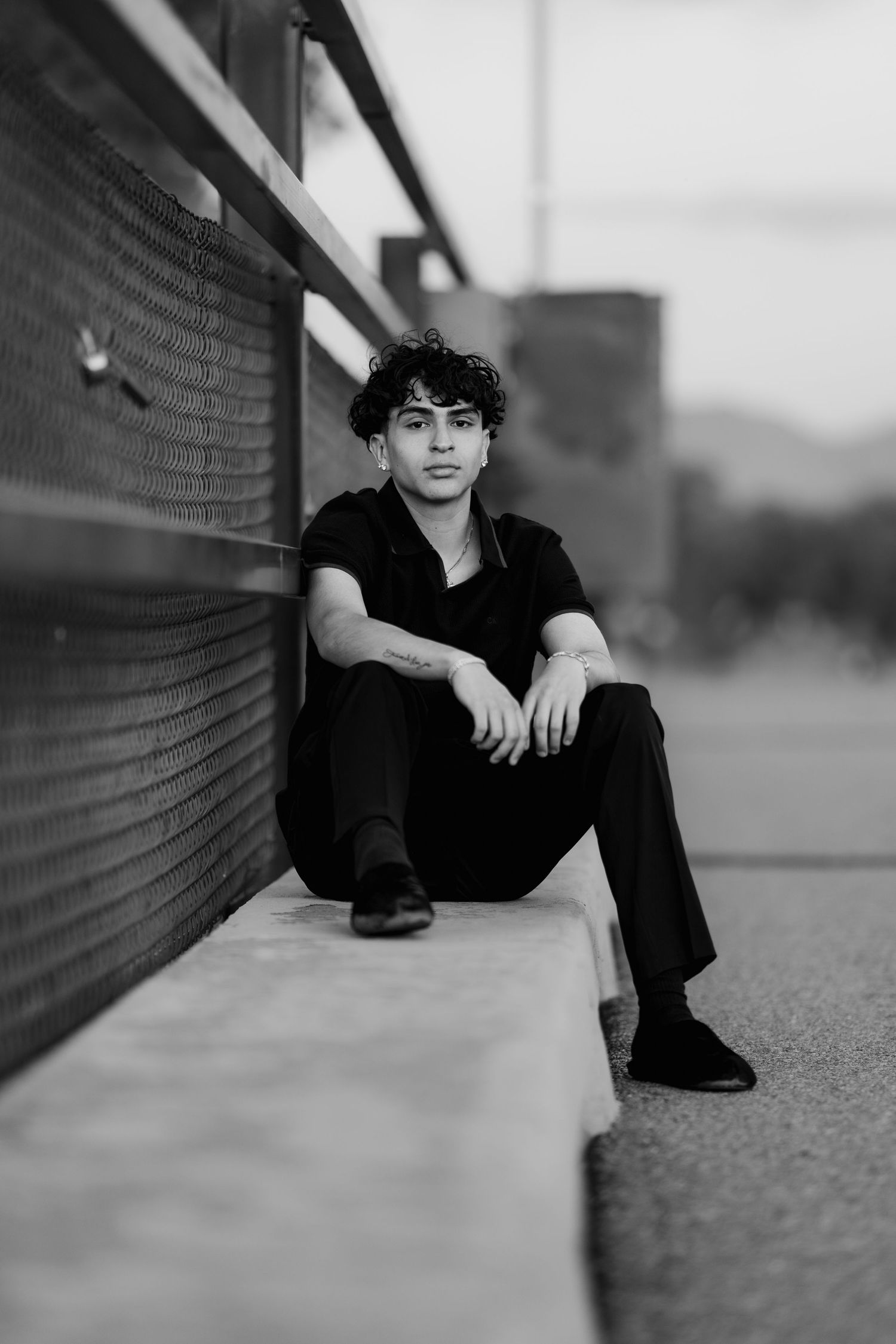 Black and white portrait of someone sitting against a wall wearing dark casual clothing in a moody urban setting.
