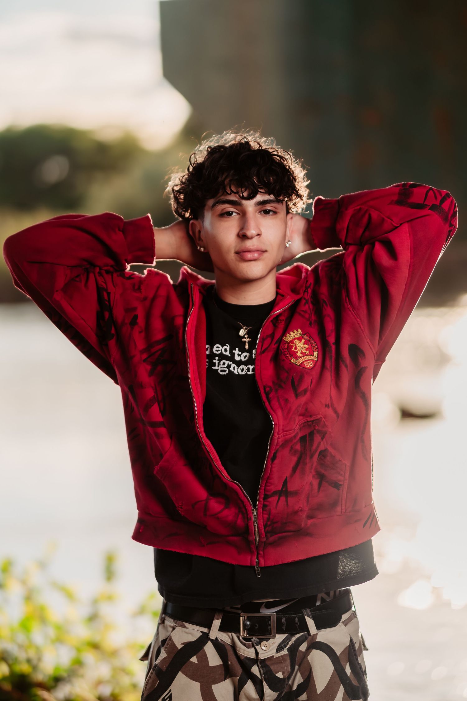 Backlit portrait showing someone in red hoodie with hands behind head against sunset.