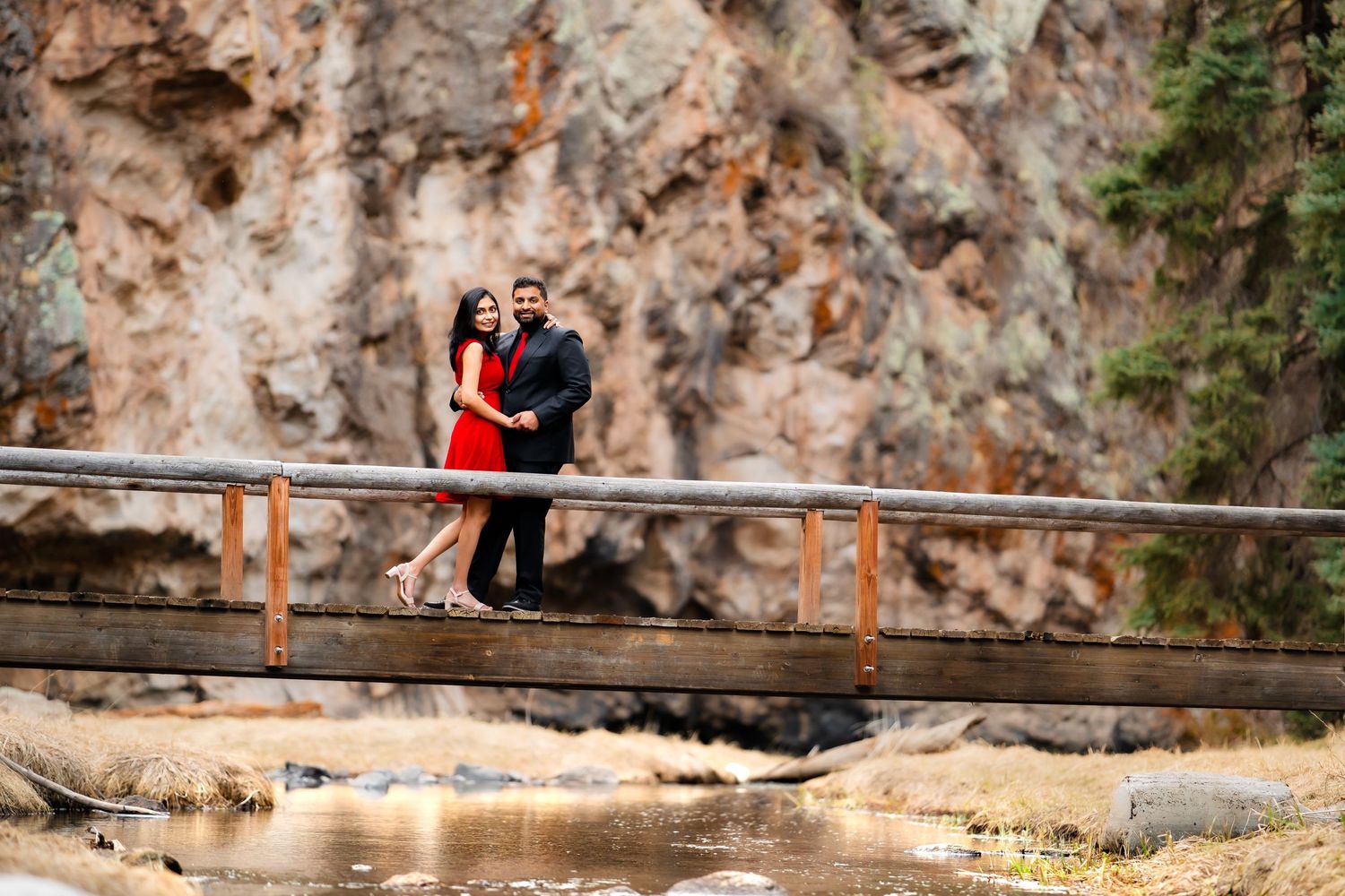 A couple in formal attire poses on a wooden bridge against dramatic rocky cliffs in nature.