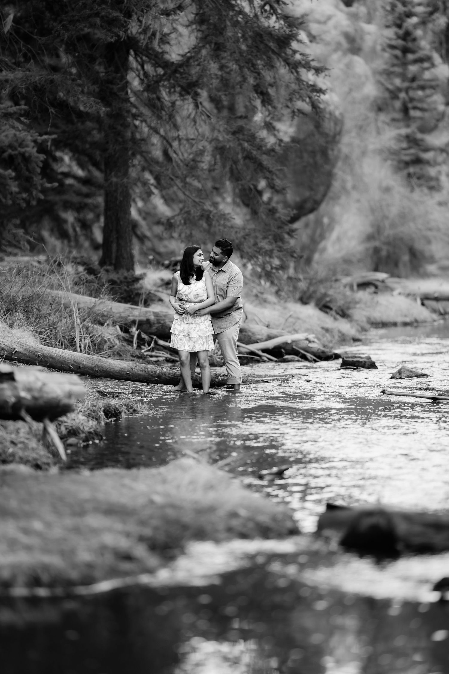 Black and white photo of couple embracing by rushing stream in forest setting.