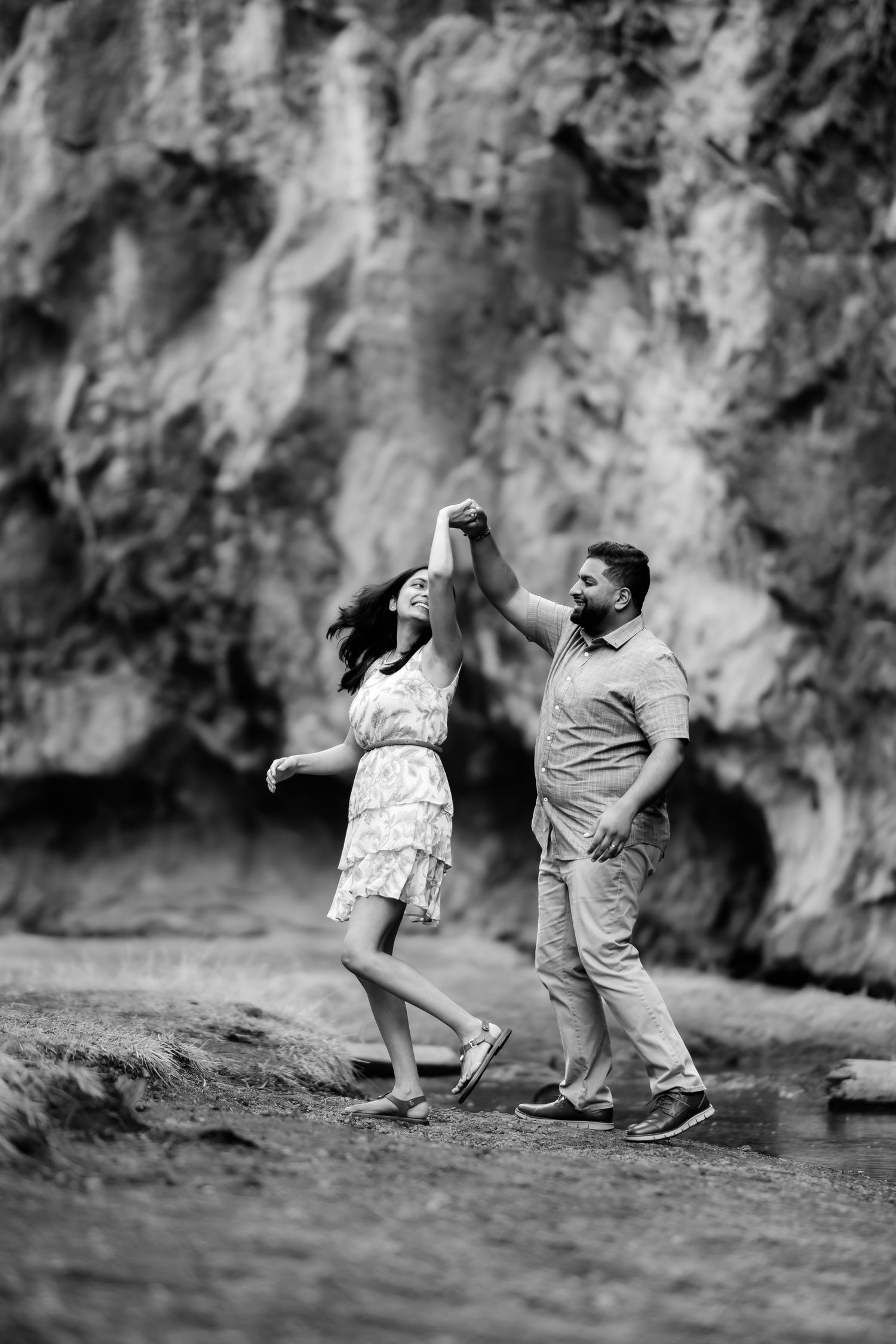 Dancing couple twirls together near cliffside in black and white outdoor photo.