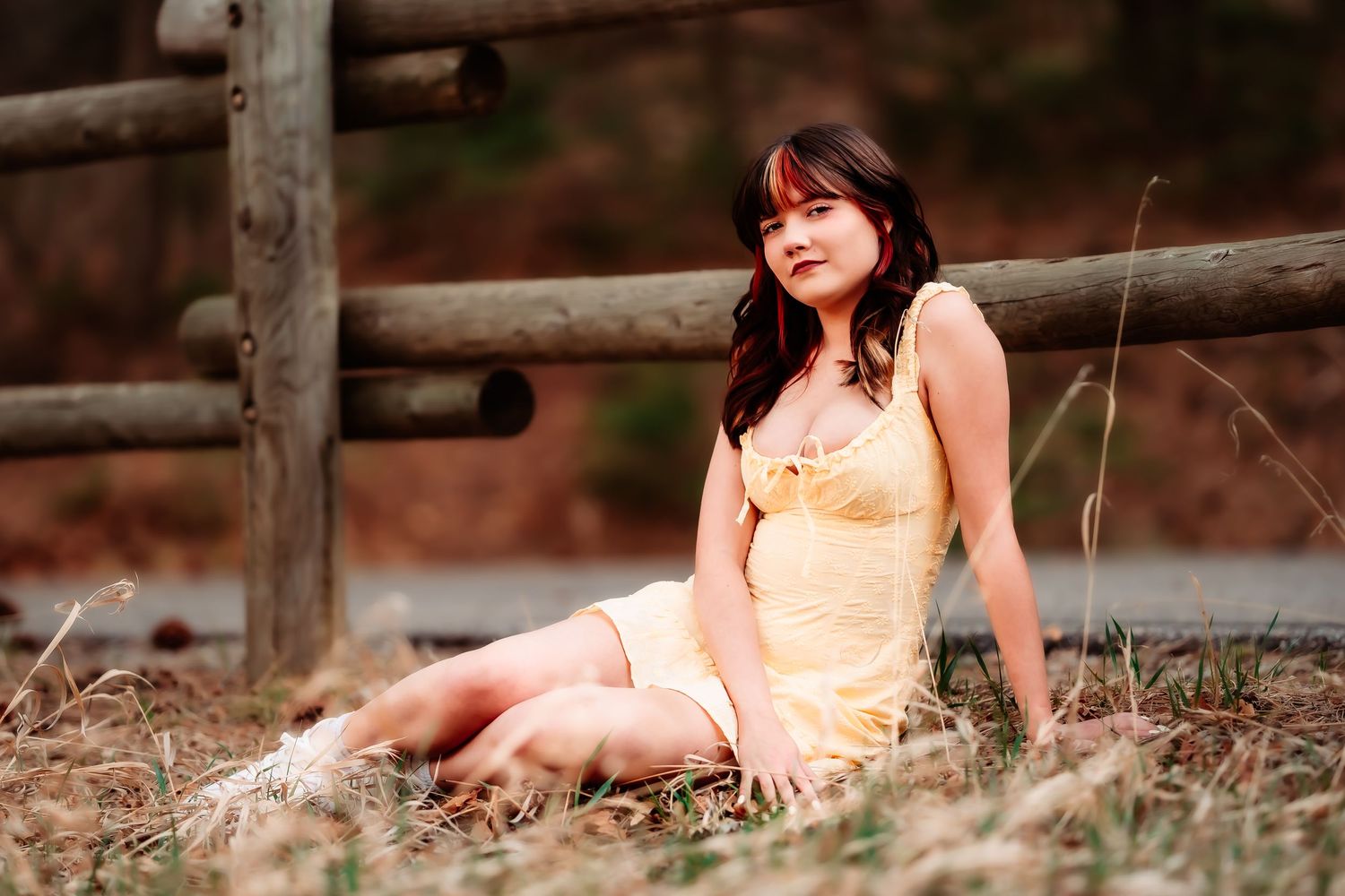 A person in a yellow sundress sits gracefully in dried grass near a rustic wooden fence.