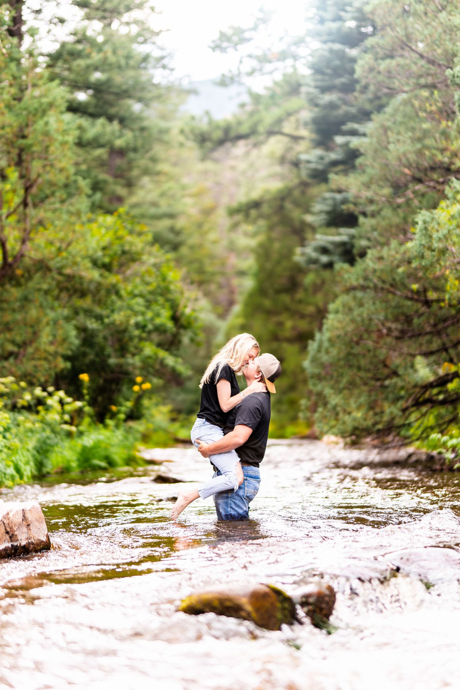 A romantic couple embraces on a rocky outcrop surrounded by lush pine forest on a bright sunny day.