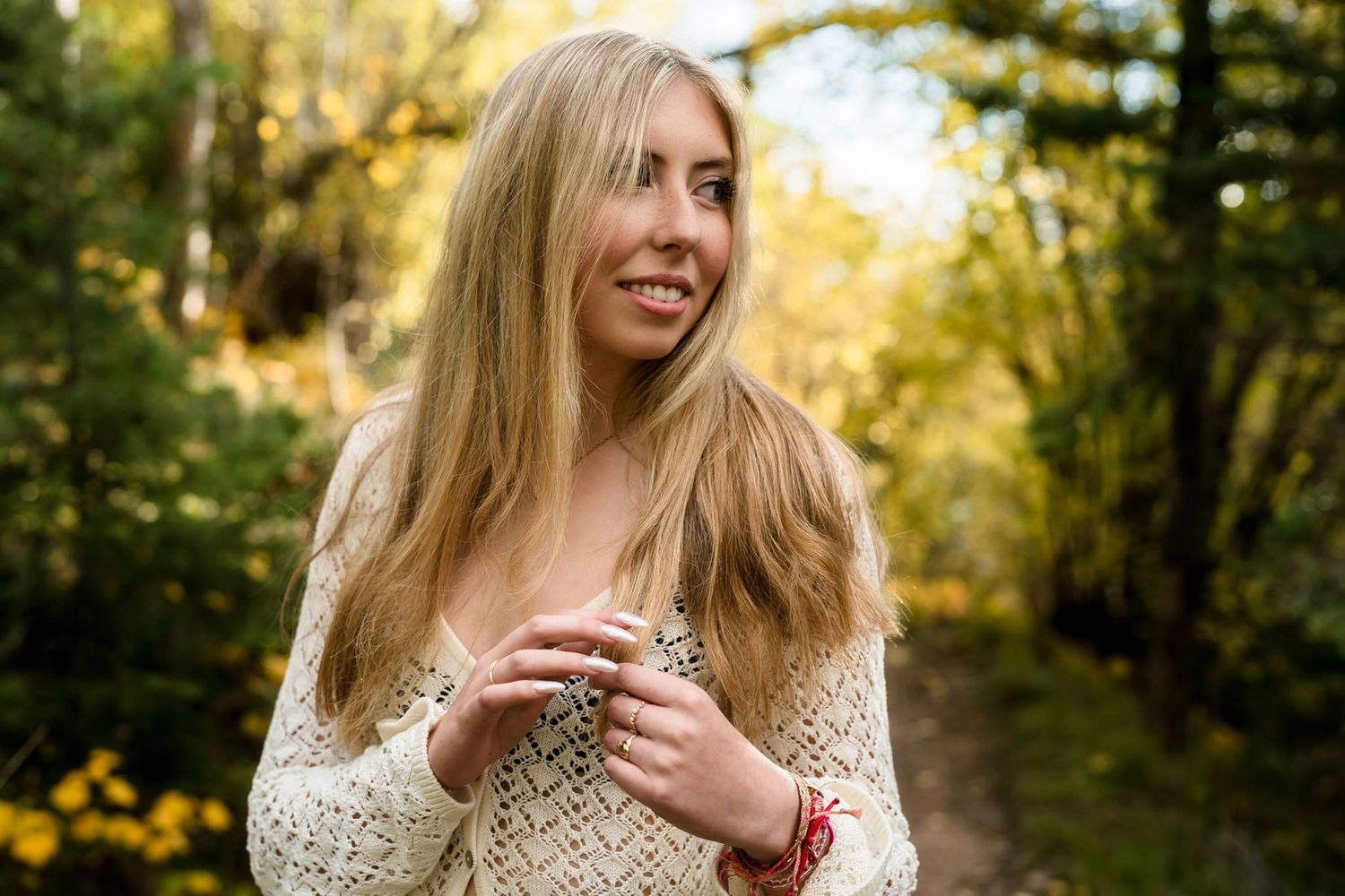 Smiling blonde woman in cream knit sweater standing on a forest path surrounded by autumn foliage.