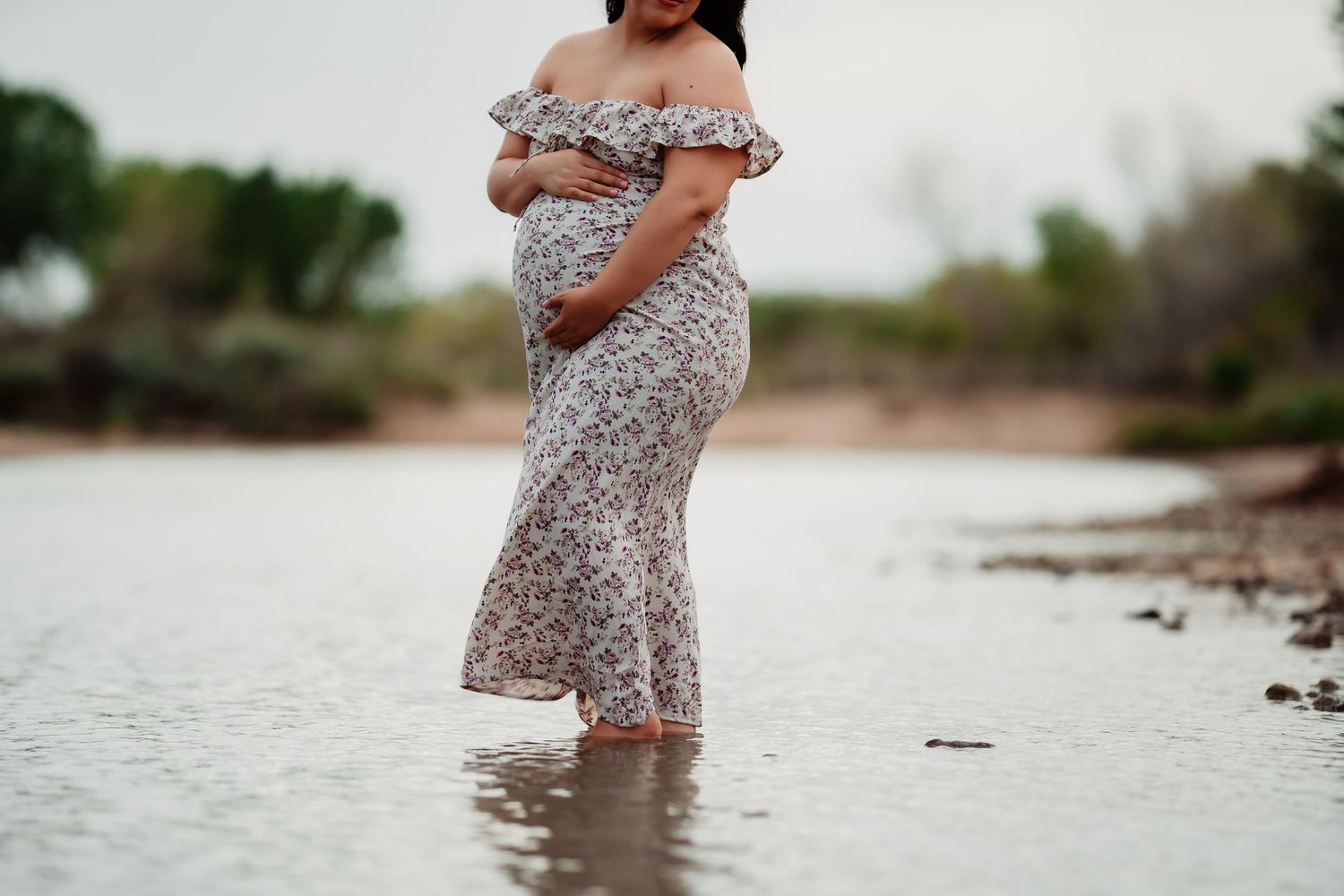 An expectant mother in a floral dress stands in shallow water at the edge of a serene lake.