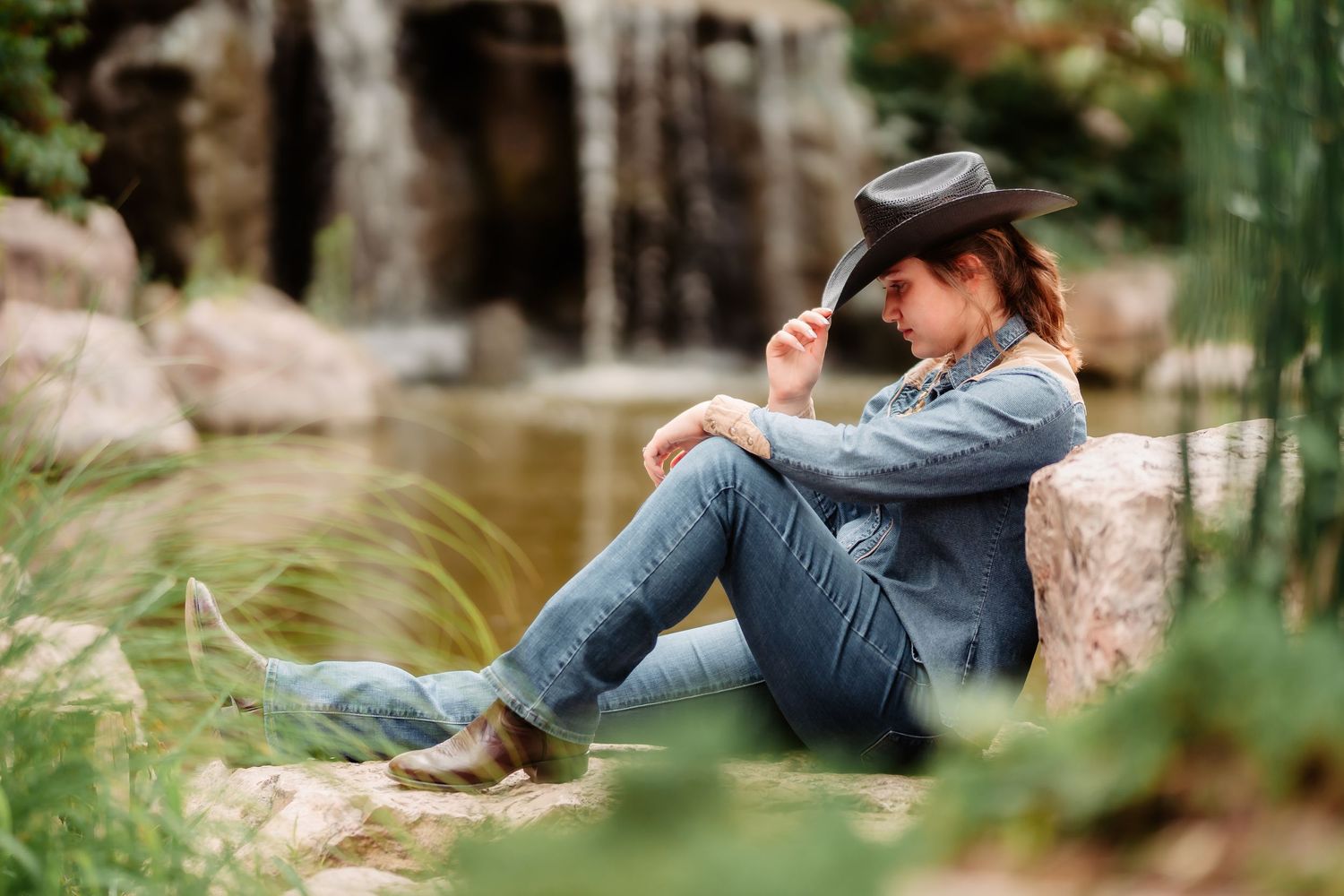 Rustic outdoor photo session in nature showing someone relaxing on rocks near a stream in casual denim attire.