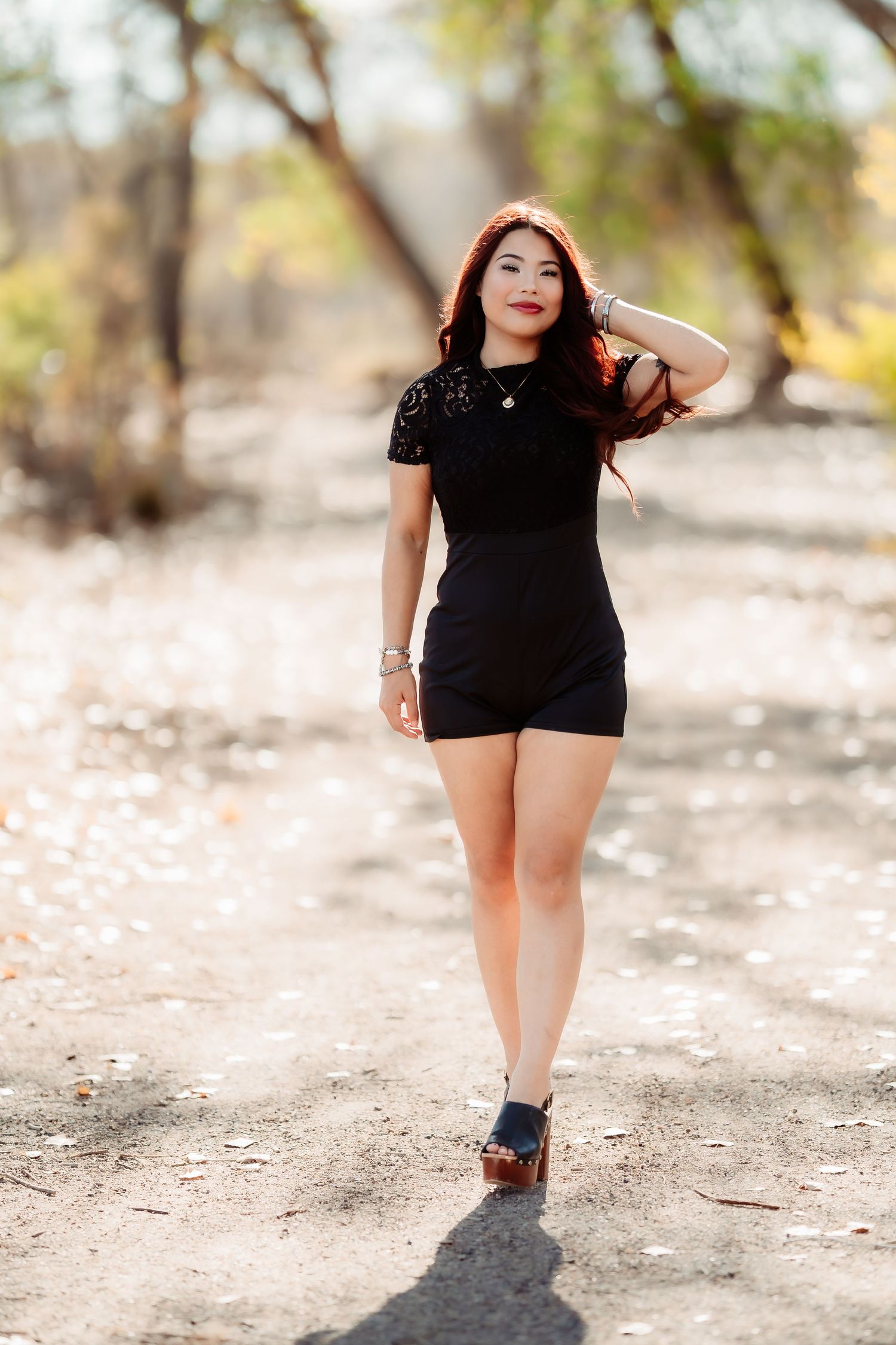 A figure in a black outfit poses outdoors on a dirt path against a natural blurred background.