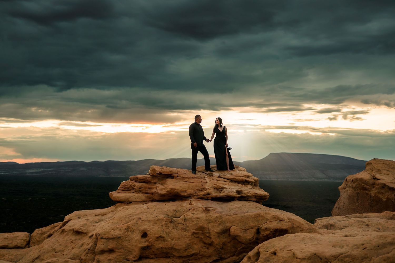 Couple holding hands on a rocky cliff at sunset with dramatic stormy skies and vast canyon landscape.