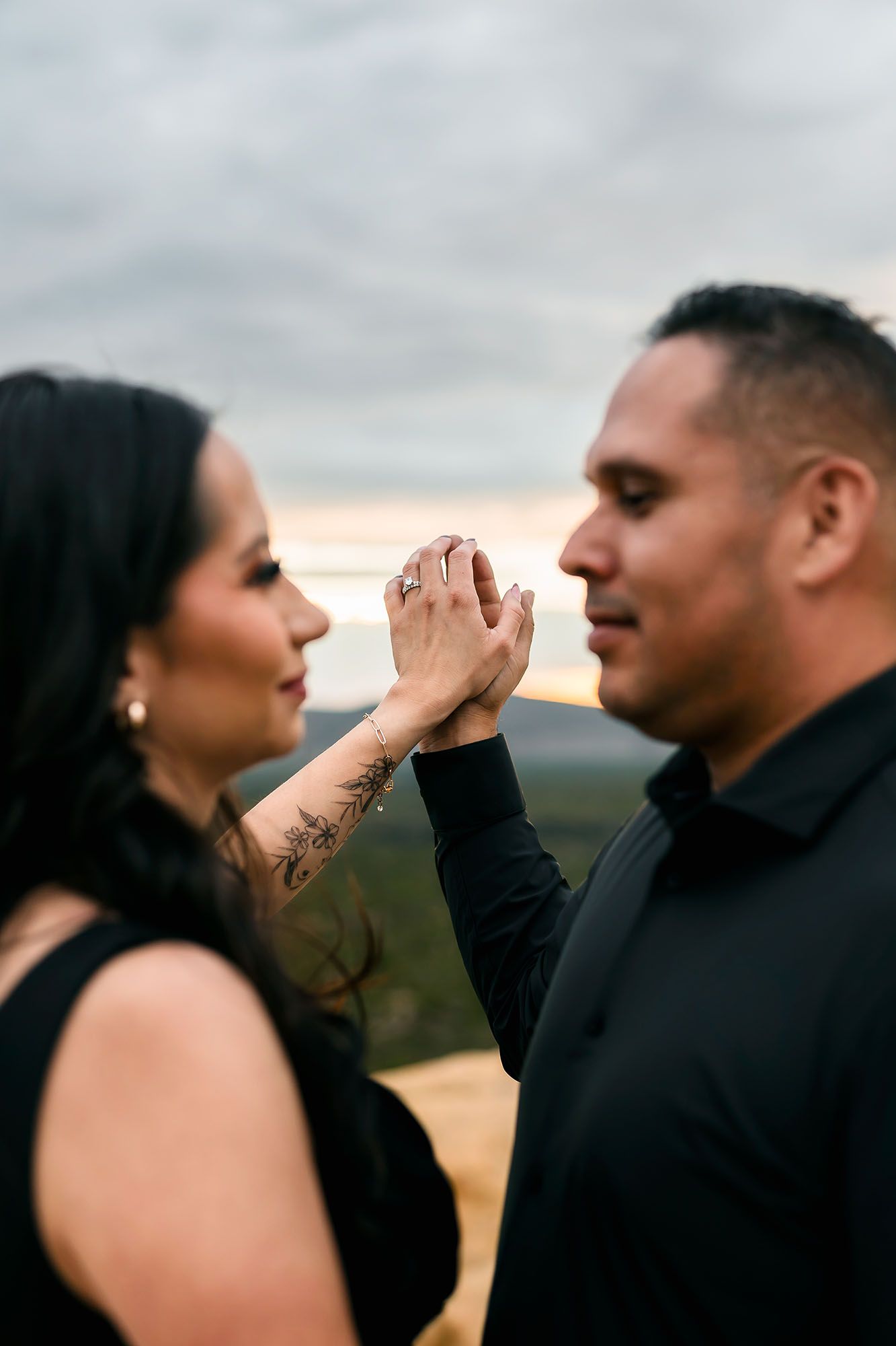 Couple in black outfits holding hands at sunset, woman displaying engagement ring with hand tattoo.