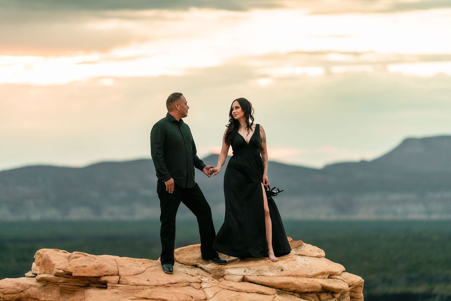 Couple in black formal wear holding hands on sandstone rock at golden hour sunset.