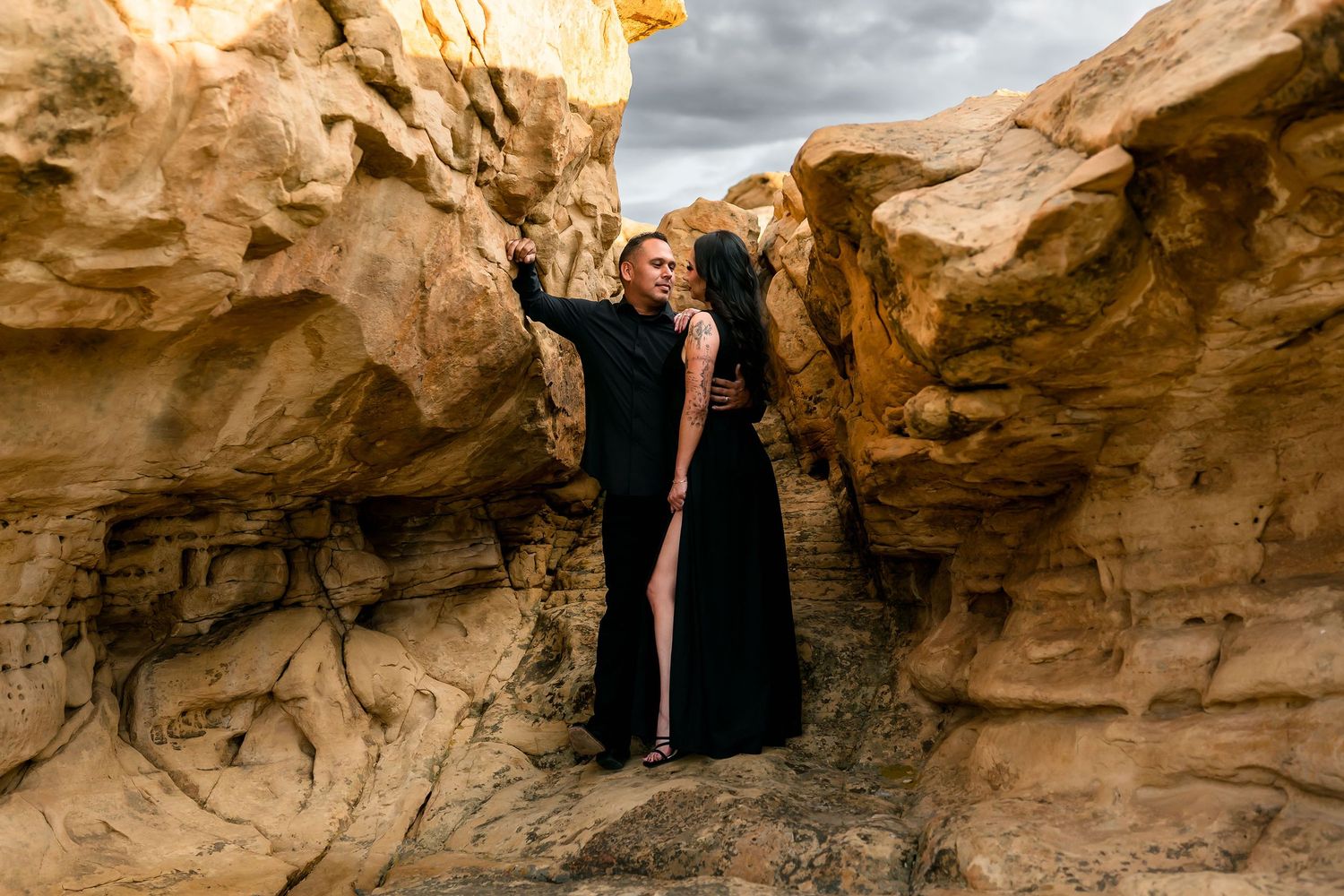Couple in black formal attire posing romantically between golden sandstone rock formations under stormy skies.