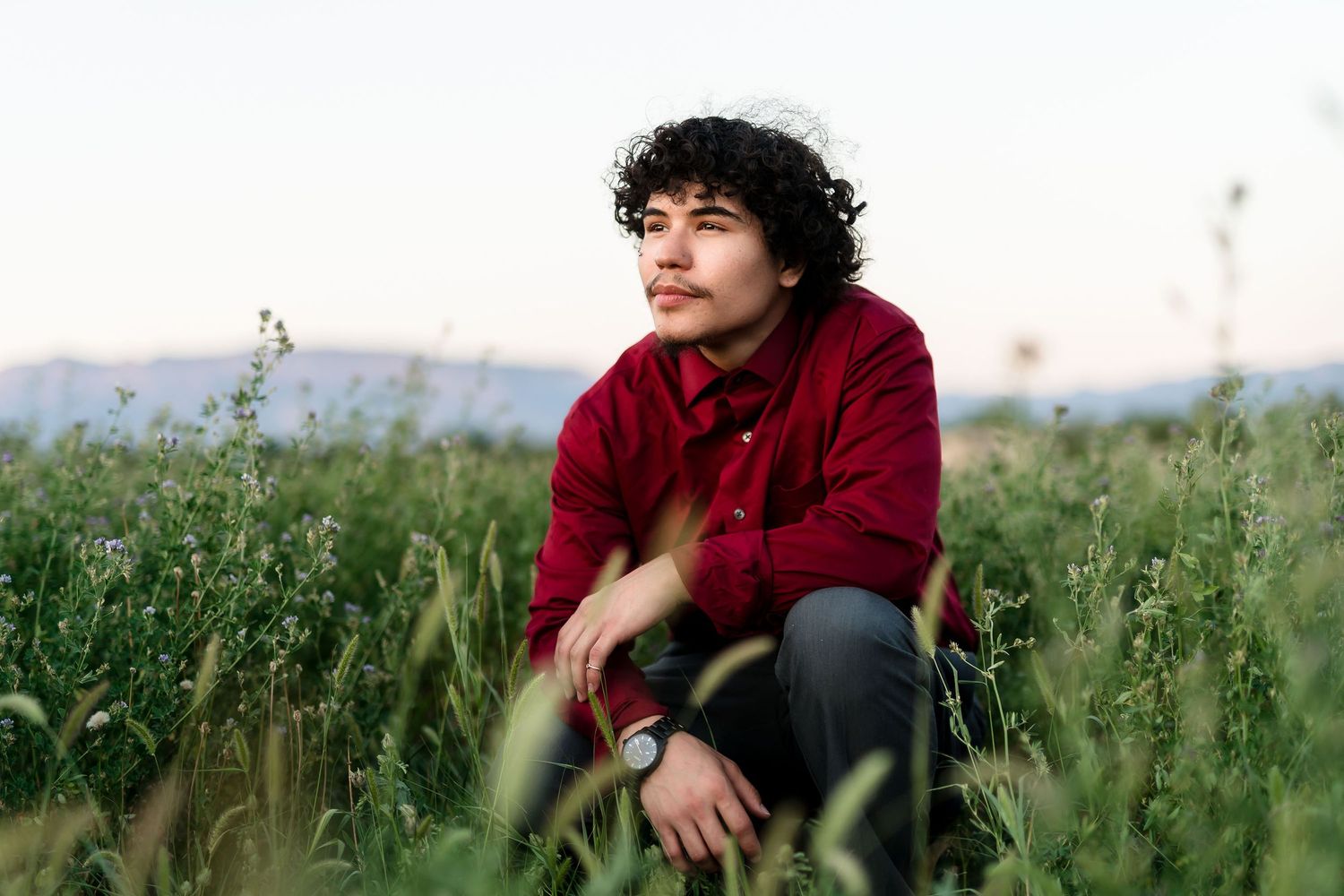 Young man with curly hair sitting in tall green field at dusk wearing a red shirt.