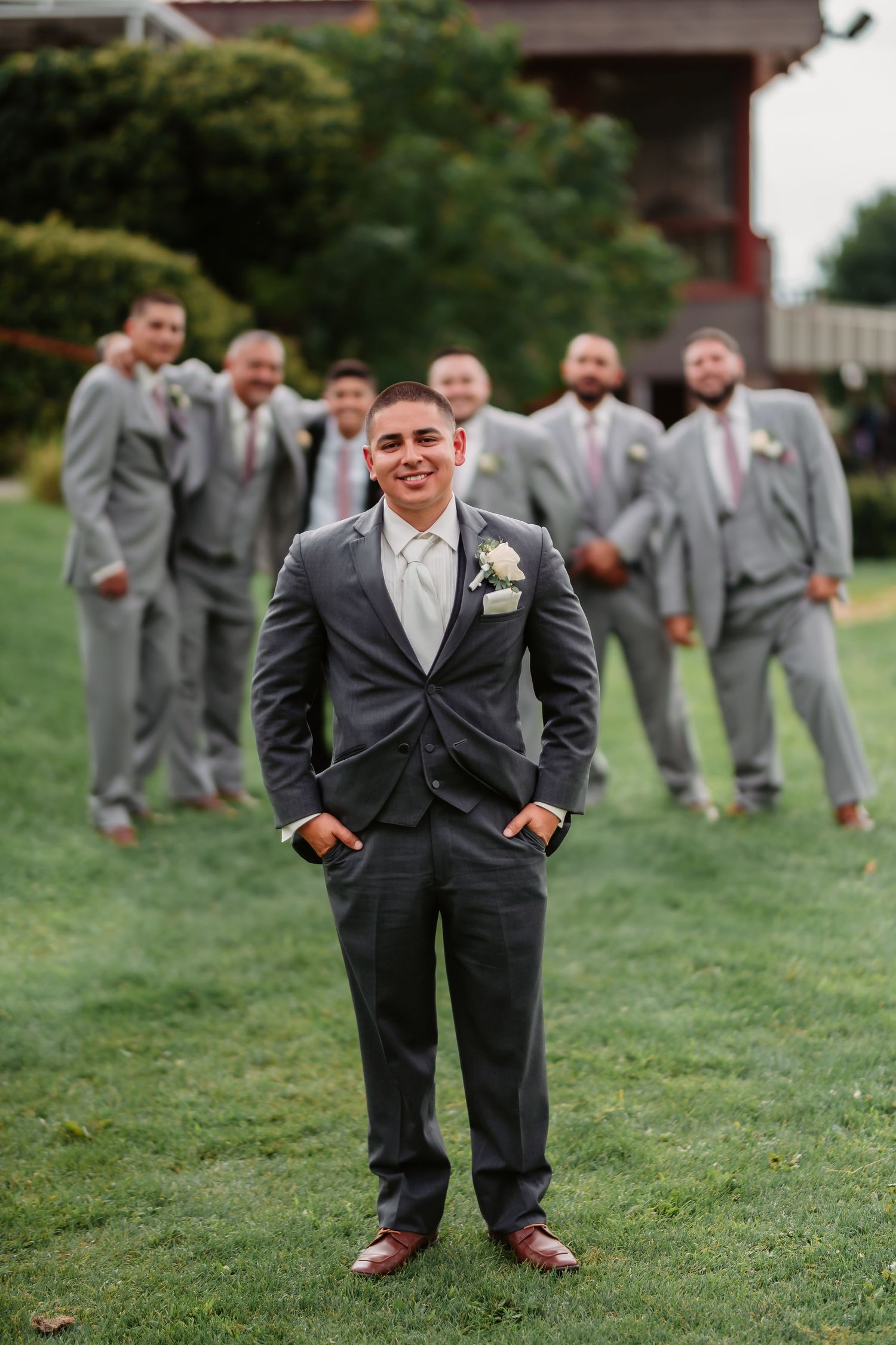 A wedding party wearing gray suits poses playfully on a grassy lawn.