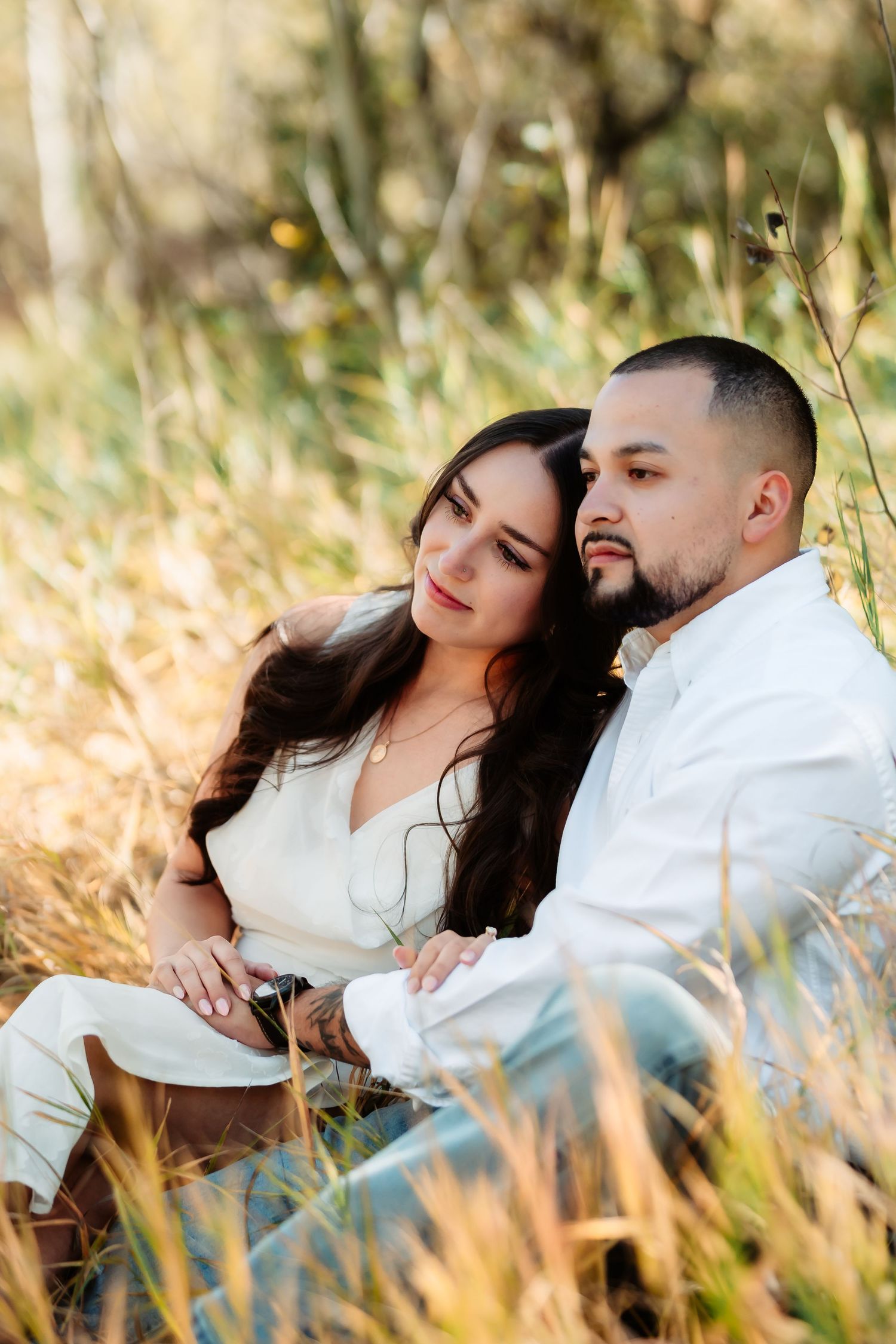 A romantic couple in white outfits sits together in a sunlit field of tall golden grass during an outdoor photo session.